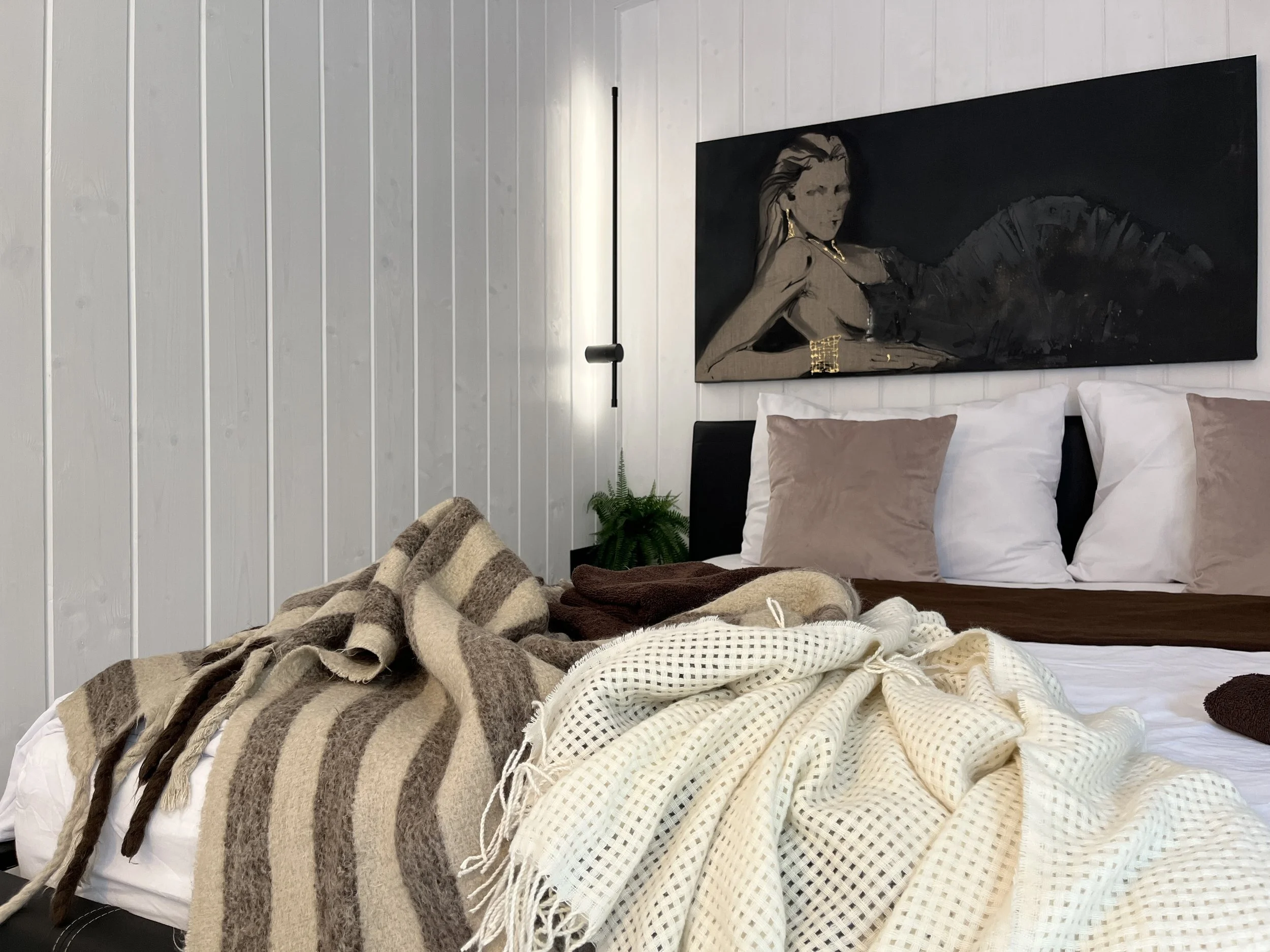 A bed with multiple blankets and pillows, a wall-mounted lamp, a piece of artwork of a woman with long hair, and a small green plant next to the bed.