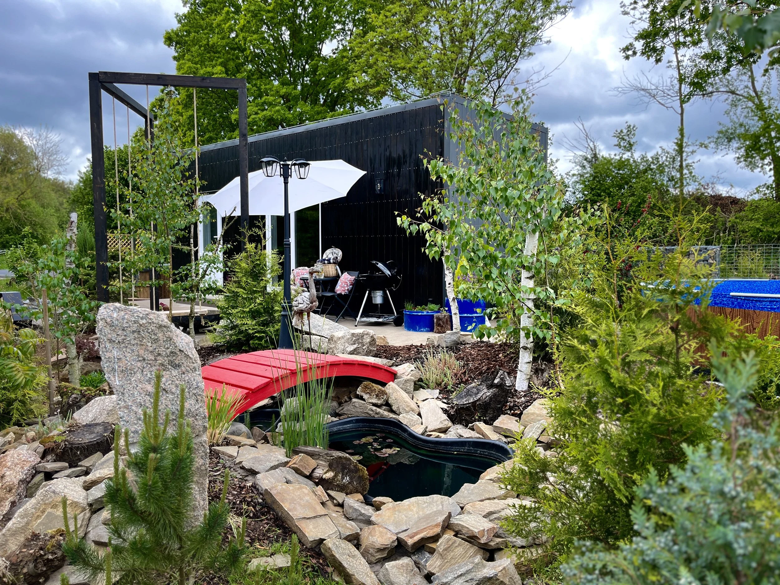 A backyard garden with a pond surrounded by rocks, a small red bridge, and various green plants and trees. There is a black building with a white umbrella and seating area in the background, along with a blue pool and outdoor furniture.