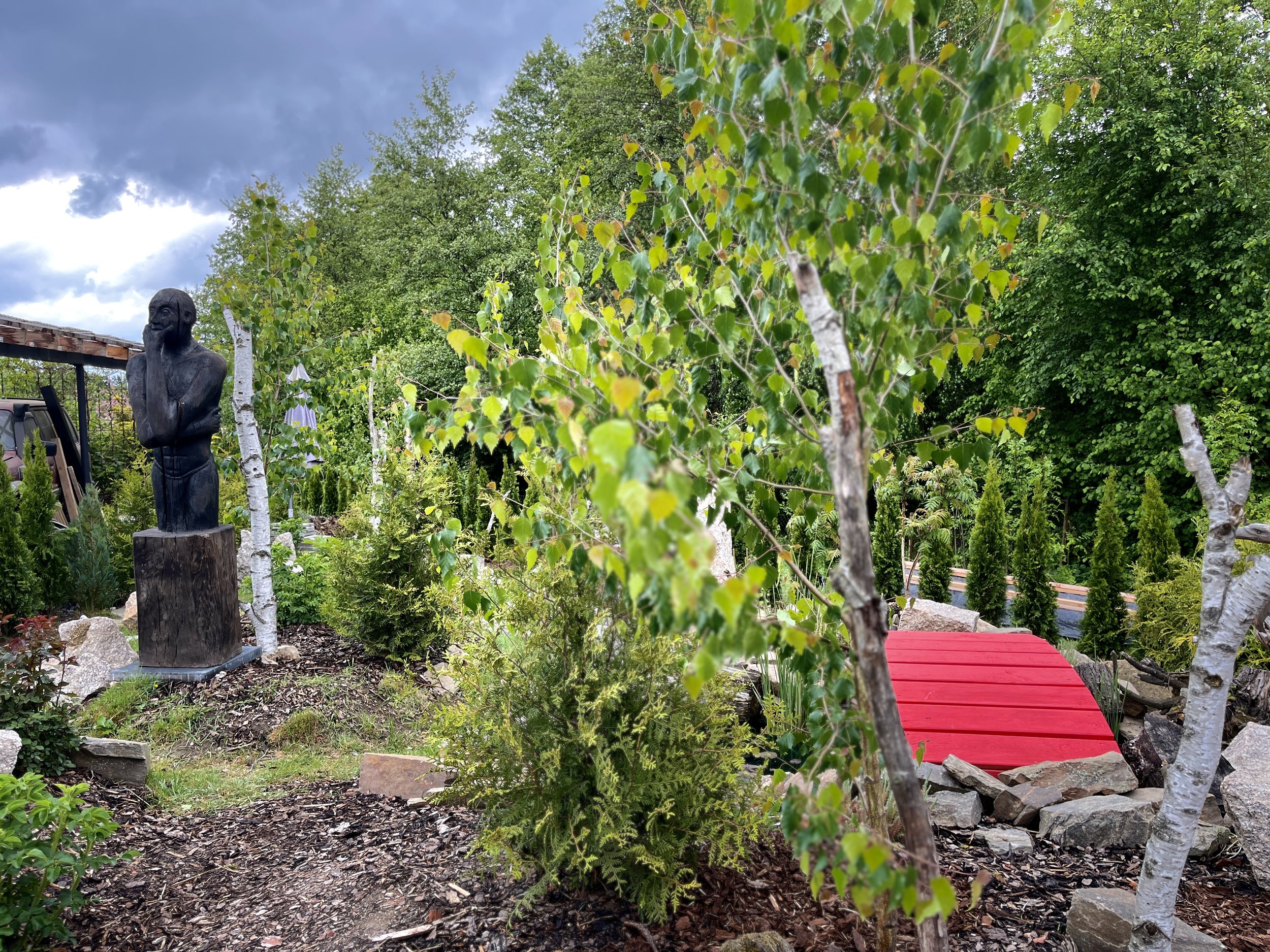A garden scene with trees, a statue of a person with a beard, and a red wooden bridge.
