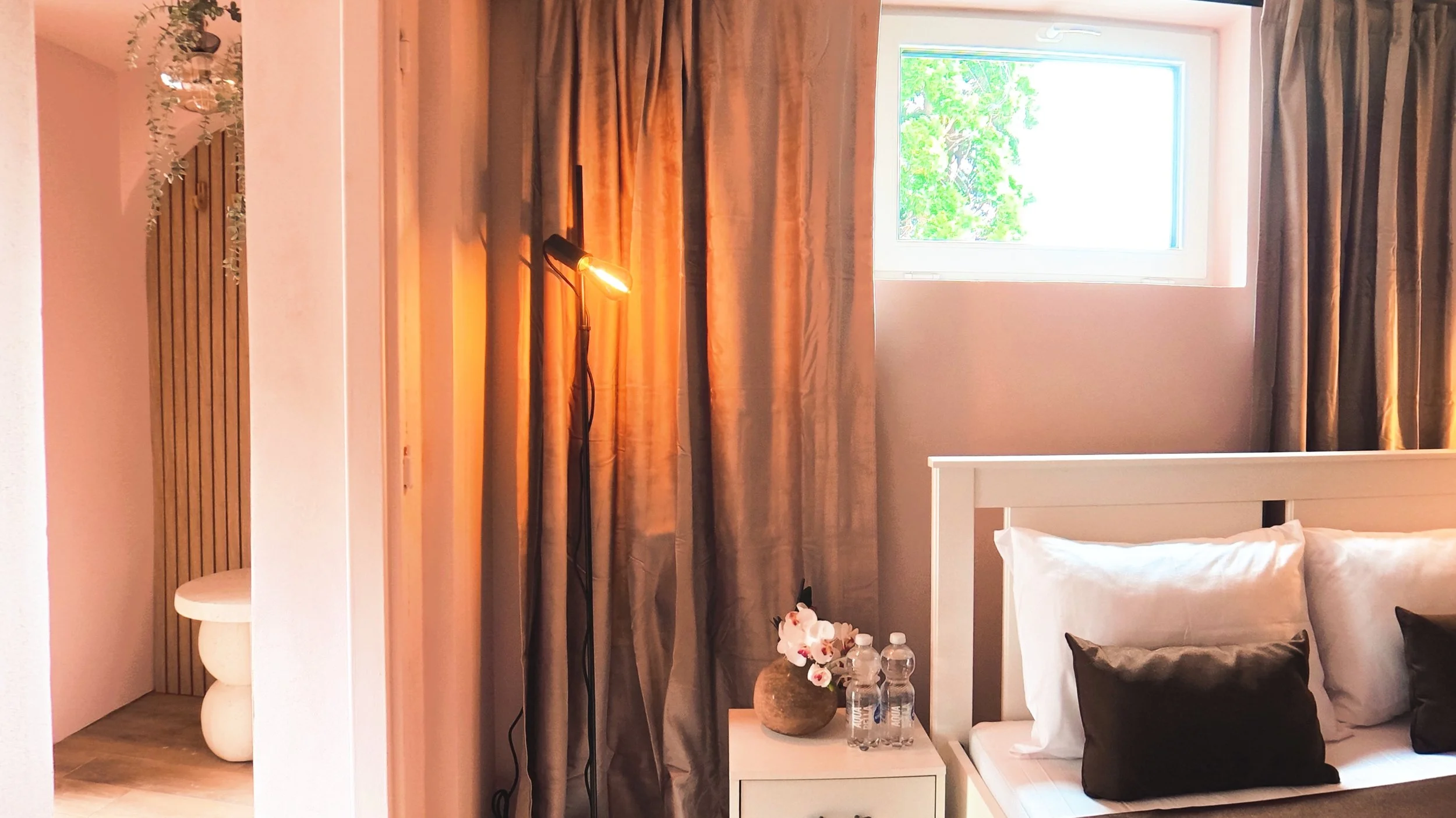Interior of a bedroom with a white bed frame, pillows, and a nightstand with water bottles and flowers, window with open blinds, curtains, a lamp, and decorative plants.