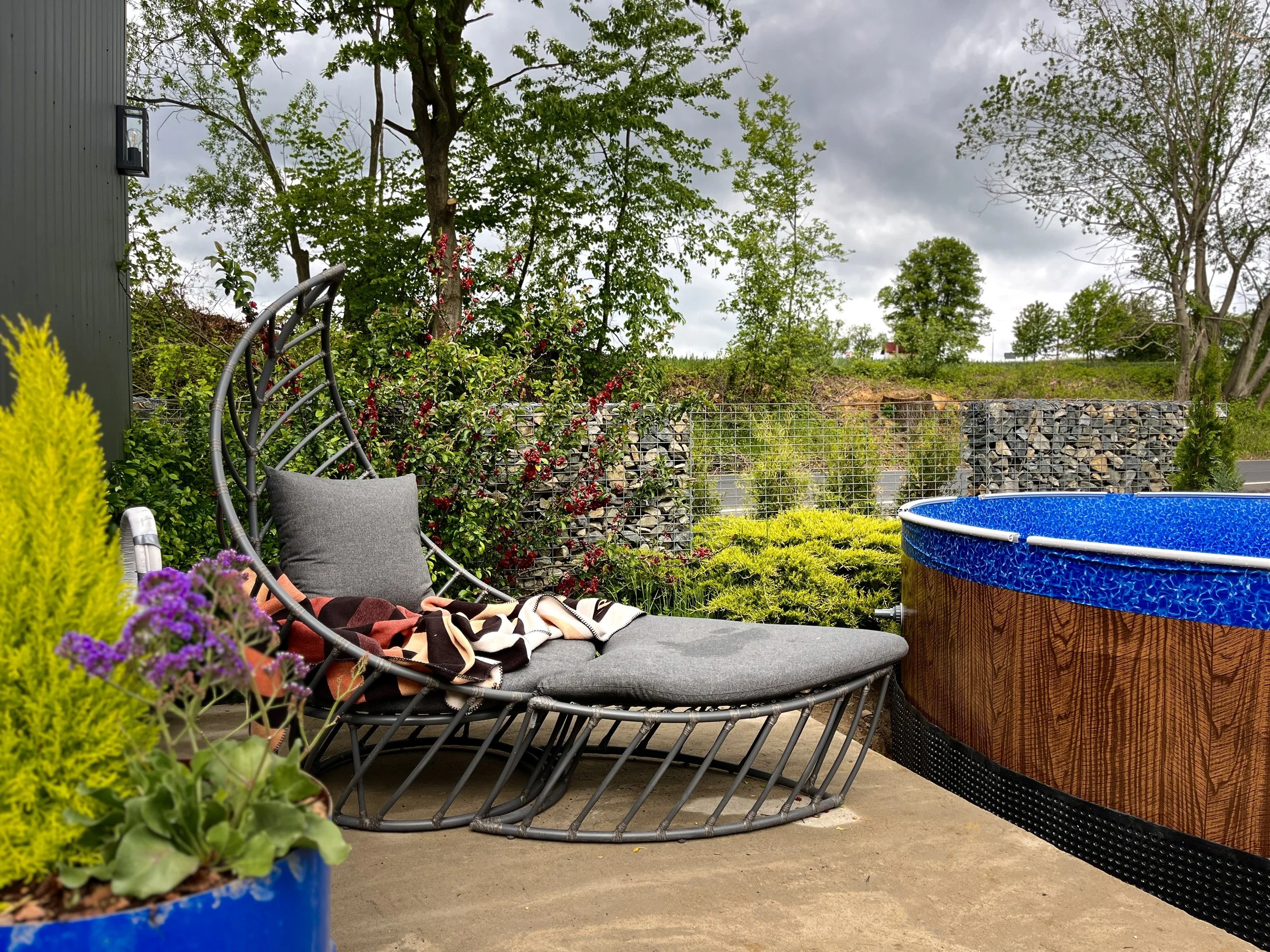A backyard patio with a cushioned lounge chair draped with a blanket, a blue round kiddie pool, a wire gabion wall, various plants, trees, and a cloudy sky overhead.