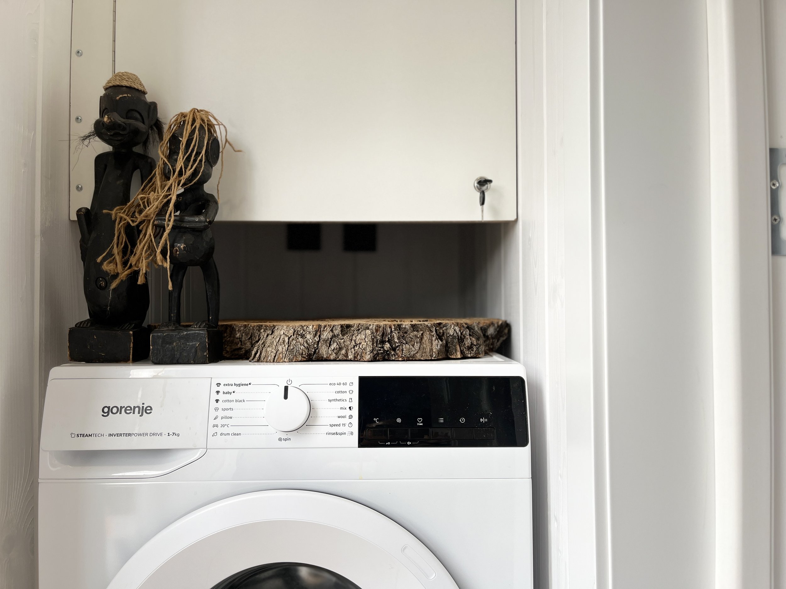 A white Gorenje washing machine with its control panel showing various wash settings and options. On top of the washing machine, there are two decorative wooden sculptures of African tribal figures standing on a small wooden platform, and a piece of cut tree trunk under them. The scene is set indoors, within a white cabinet or small closet.