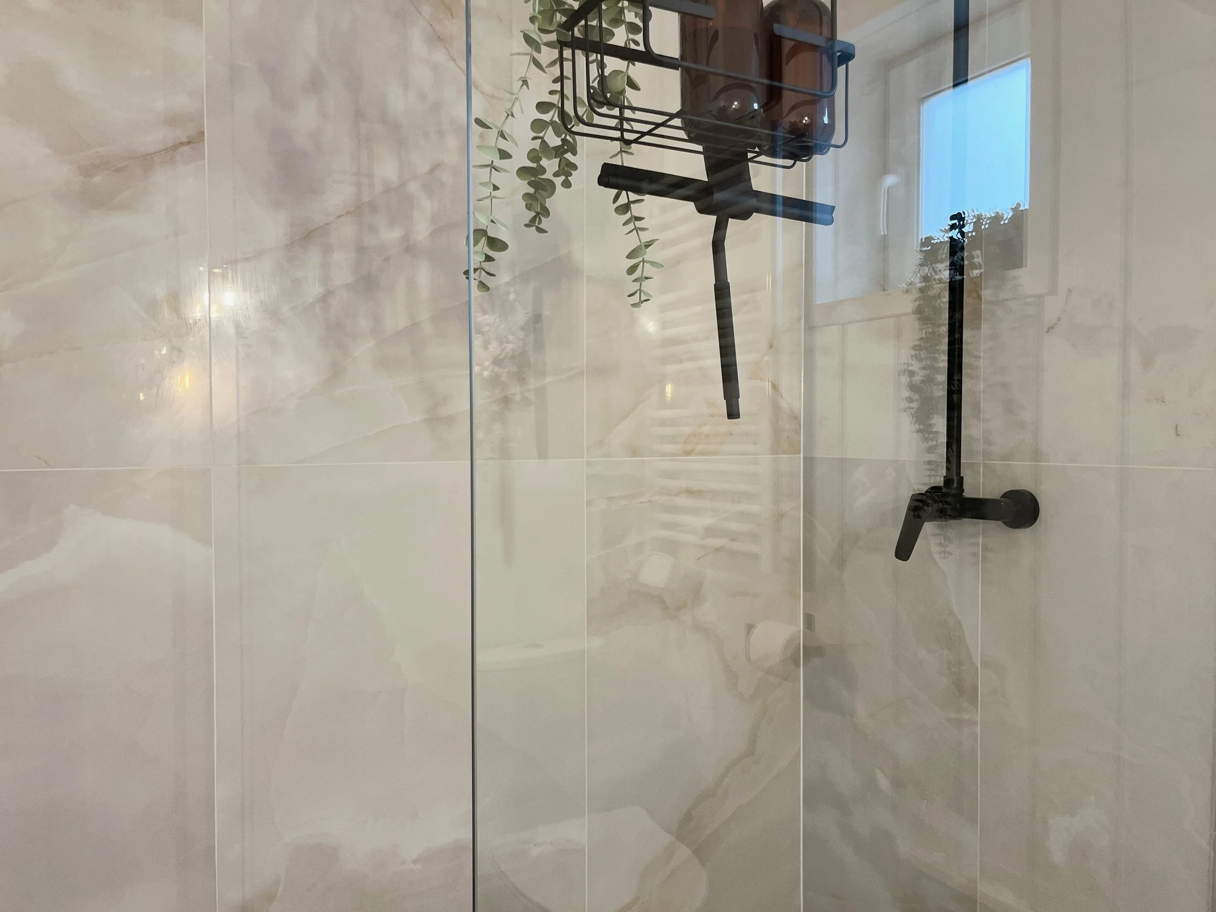 A hand truck with a leafy plant, a tray with small potted plants, and a brown glass bottle inside a glass room.