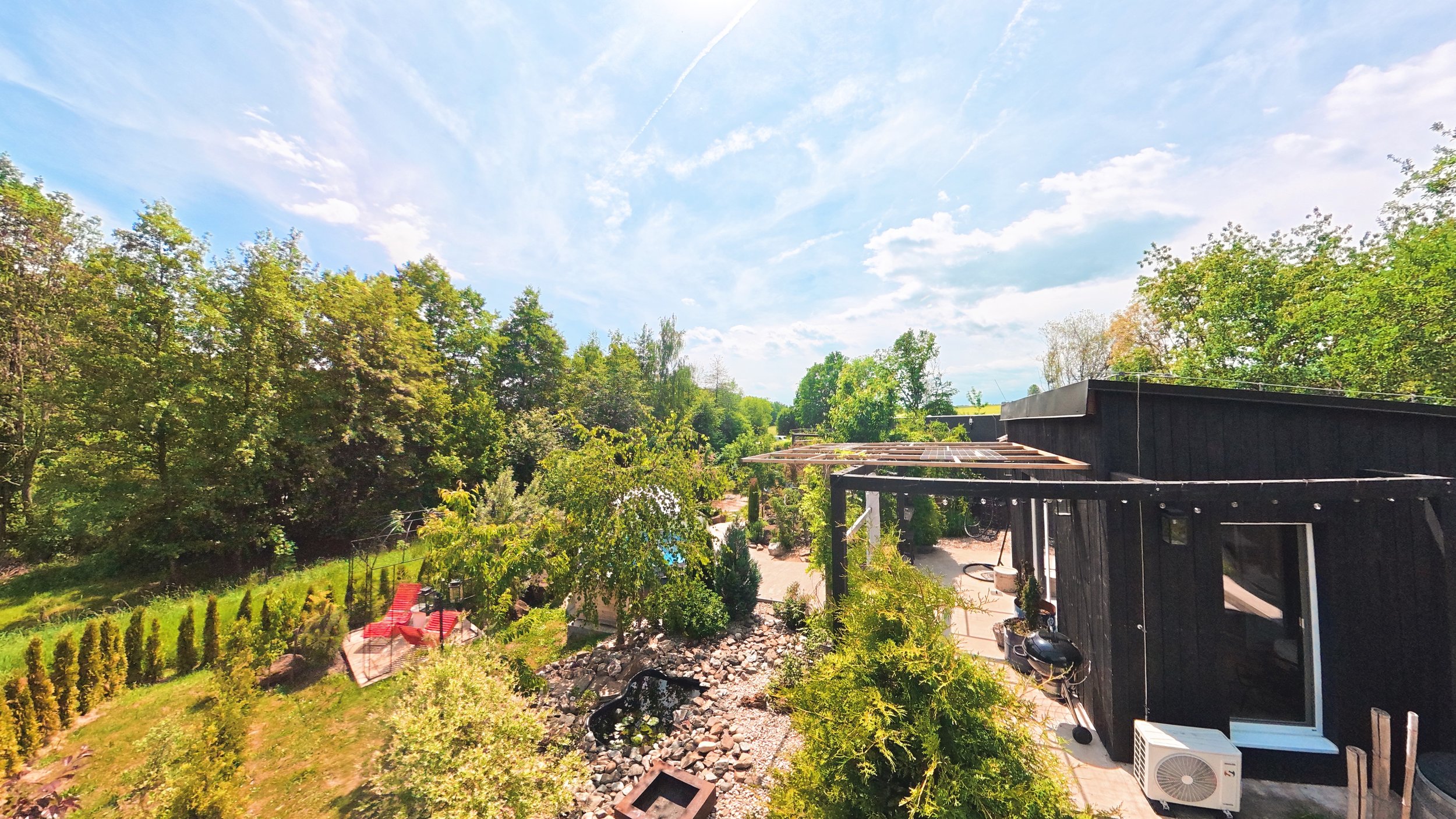 Late spring or early summer view of a backyard with lush green trees, a black modern house, patio furniture, and a small pond, under a partly cloudy blue sky.