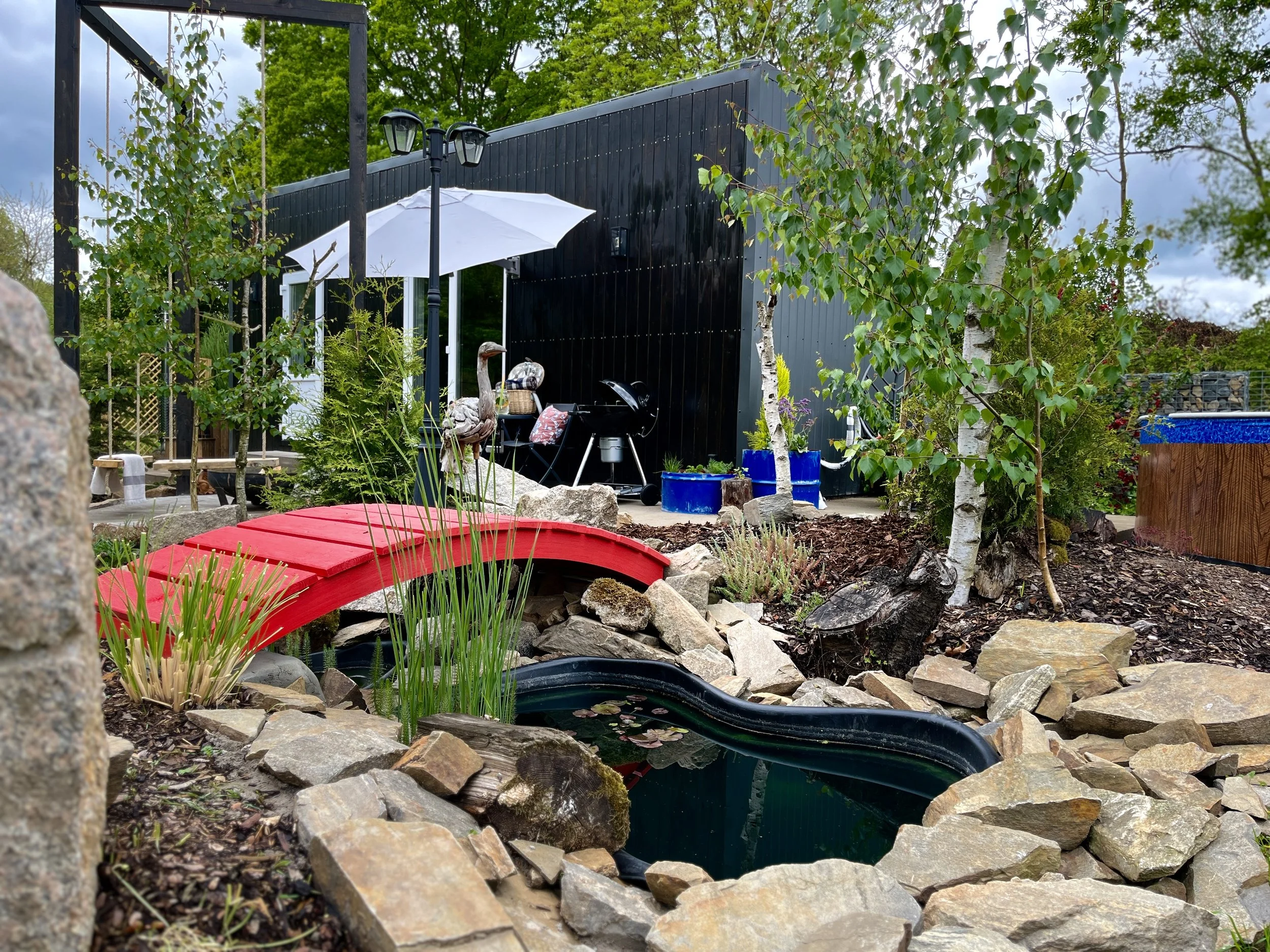 A garden area with a small pond surrounded by rocks, various plants, and trees. There is a red bridge in the foreground, a black shed or structure in the background, and a white patio umbrella near a barbecue grill.