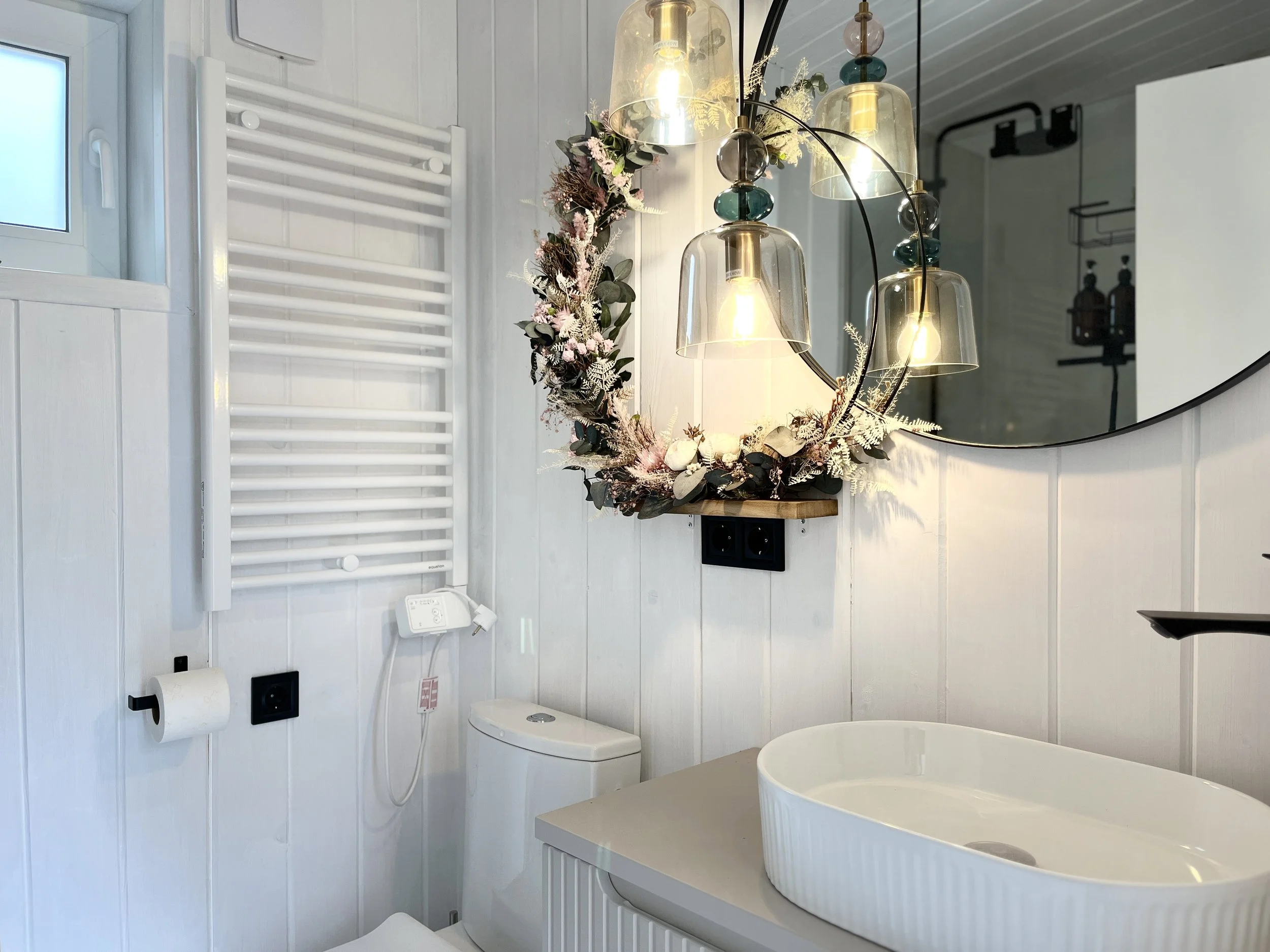 A bathroom featuring a heated towel rail on a white paneled wall, a window, a toilet, a circular vessel sink on a vanity, and a modern circular mirror. There is a decorative floral arrangement and hanging light fixtures above the sink, with electrical outlets and a plug visible on the wall.