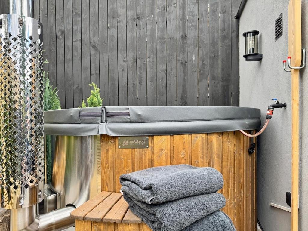 Hot tub on a deck with folded gray towels on a wooden bench, against a black wood fence, and outdoor wall with a light fixture.