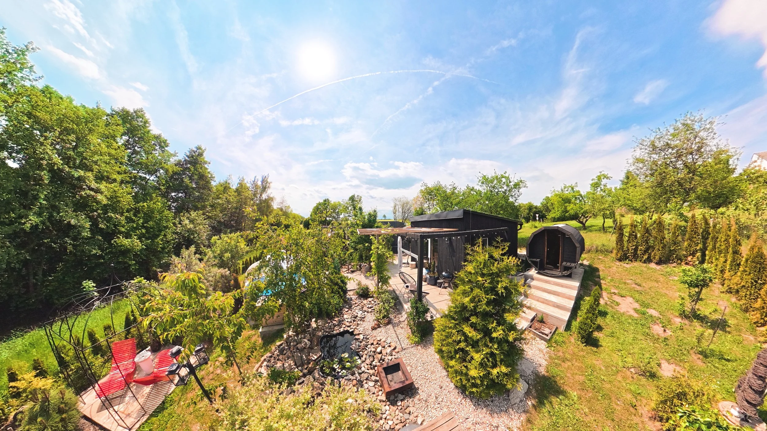 A backyard with a swimming pool, surrounded by trees and outdoor furniture, with a black shed, lounge chair, and a small rounded structure, under a bright sky with some clouds.