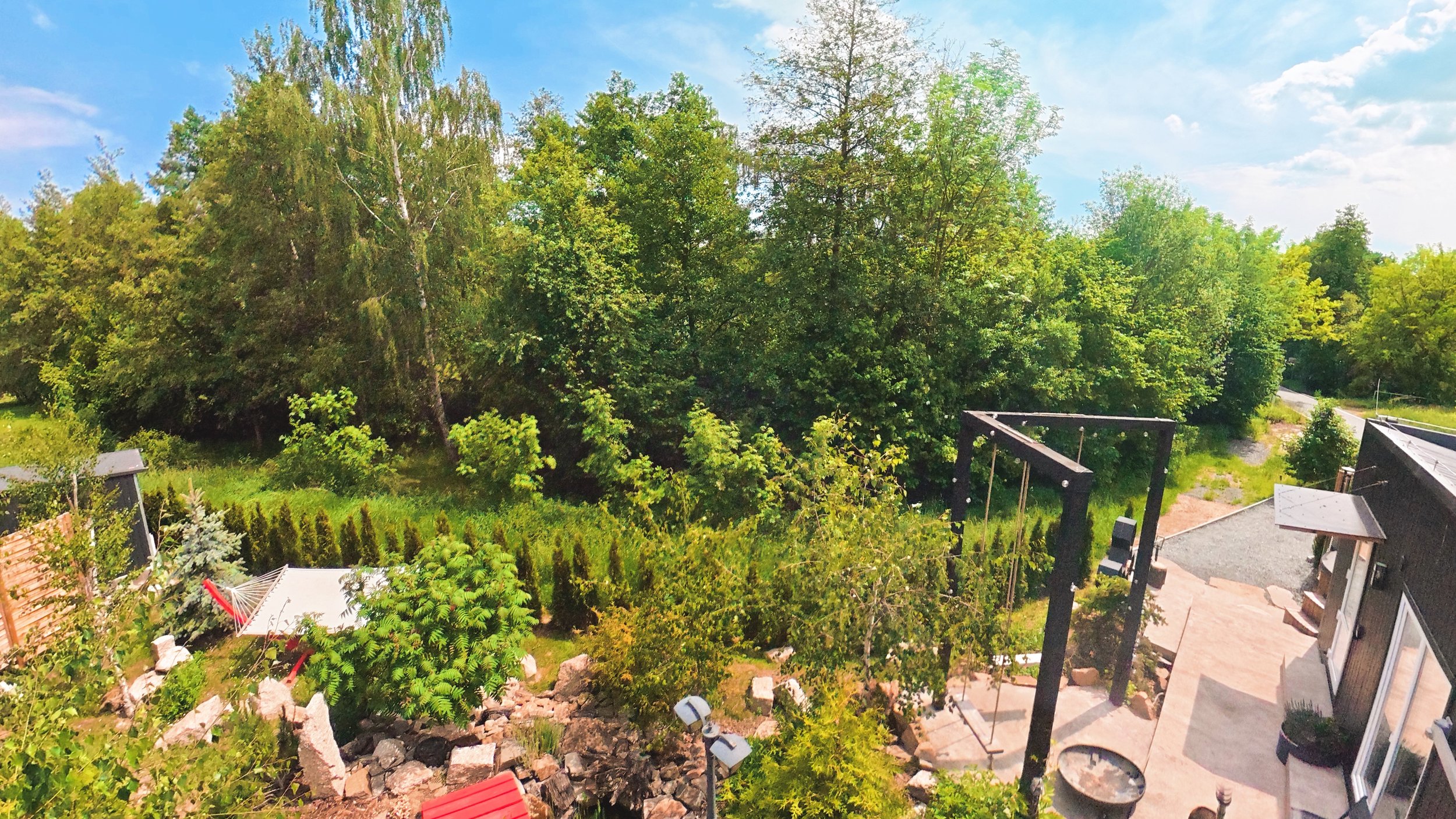 A backyard view with trees, a patio, and a small shed with a red roof. There is a garden with plants, rocks, and decorative lights, and a small rock-lined stream or drainage area. The sky is blue with some clouds.