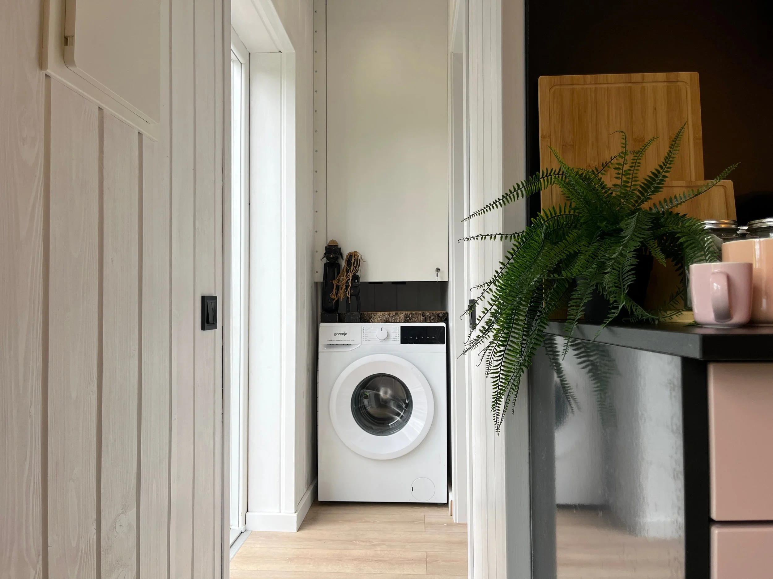 A laundry area with white wooden walls and flooring, containing a front-loading washing machine, a bamboo cutting board, potted fern, pink mugs, and jars, with a black and gold decorative item on a shelf in the background.