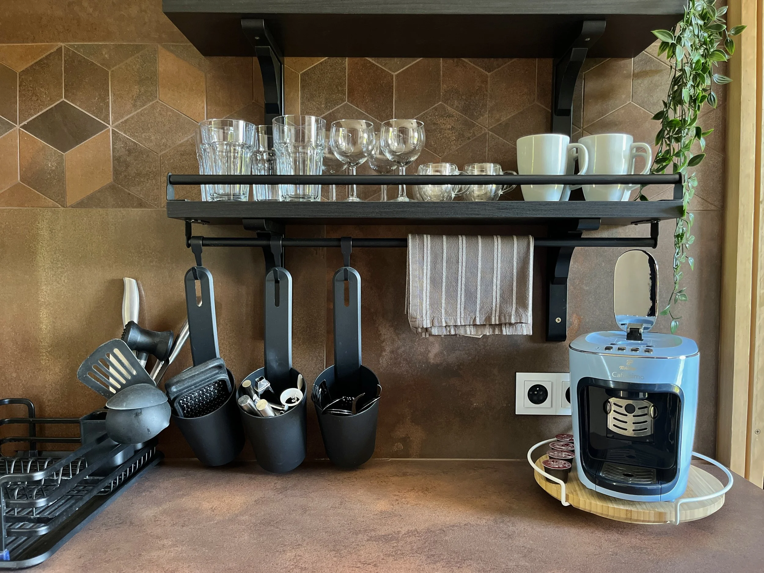 A kitchen shelf holding drinking glasses, coffee mugs, bowls, and a coffee machine, with kitchen utensils stored below and a plant on the right.