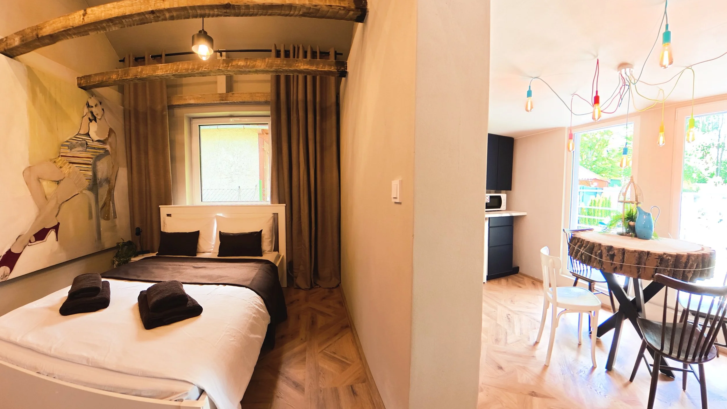A cozy bedroom with a white bed, black pillows, and folded towels, adjacent to a kitchen and dining area with a round wood table, colorful pendant lights, and large windows showing greenery outside.