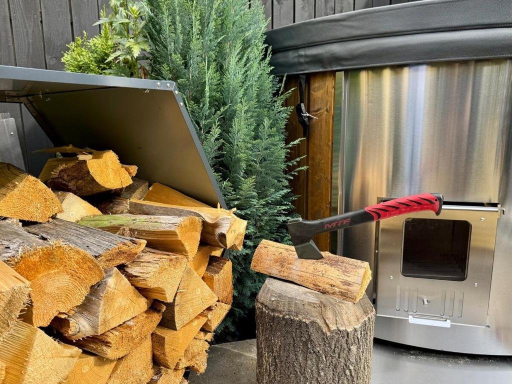Firewood stacked next to a green pine tree and a stainless steel outdoor pizza oven, with an axe embedded in a small log.