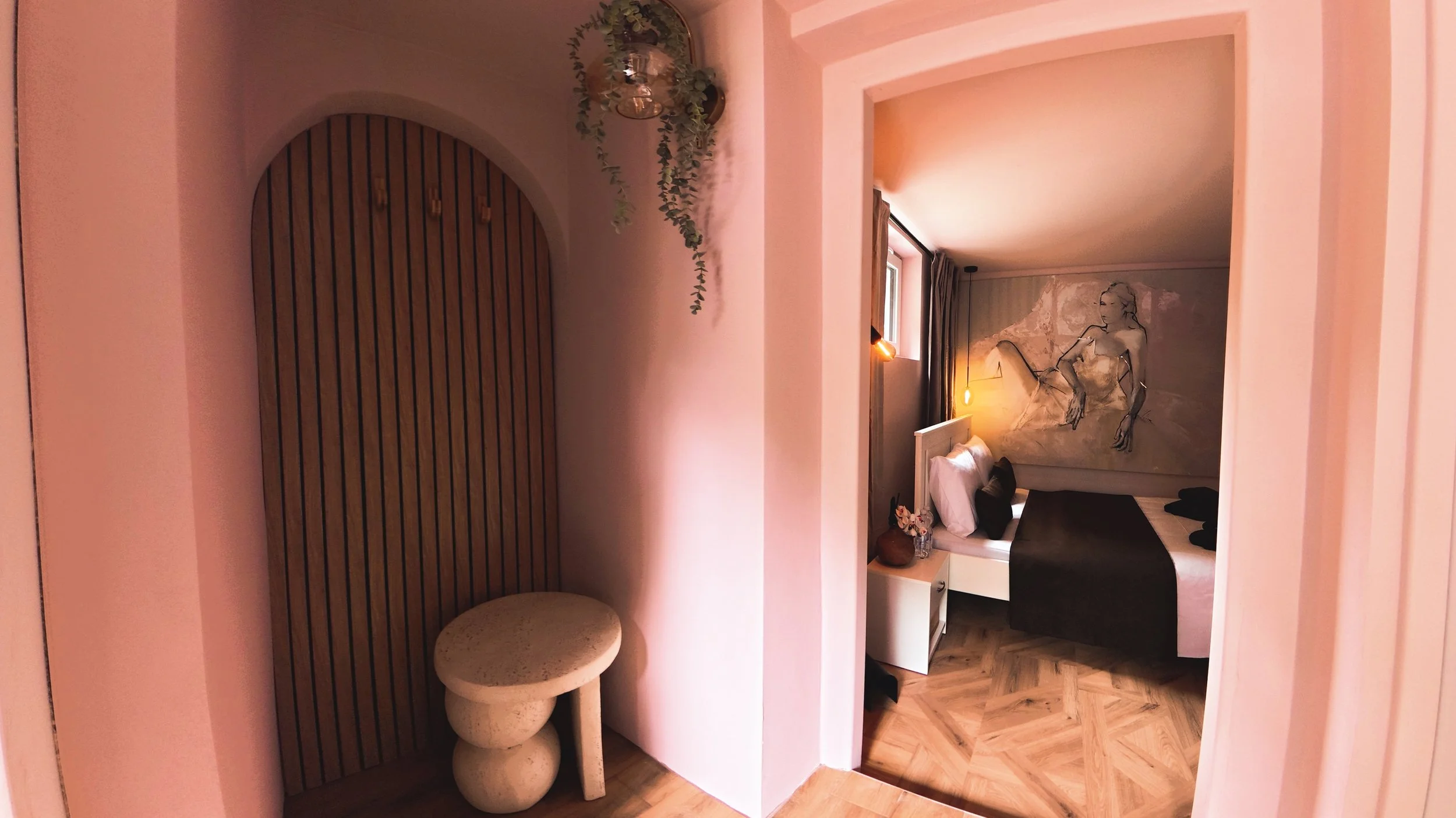 A view into a cozy bedroom with a large abstract wall art of a woman, a bed with dark bedding and pillows, a bedside table with flowers, and a window with curtains. The room has soft lighting and wooden flooring.