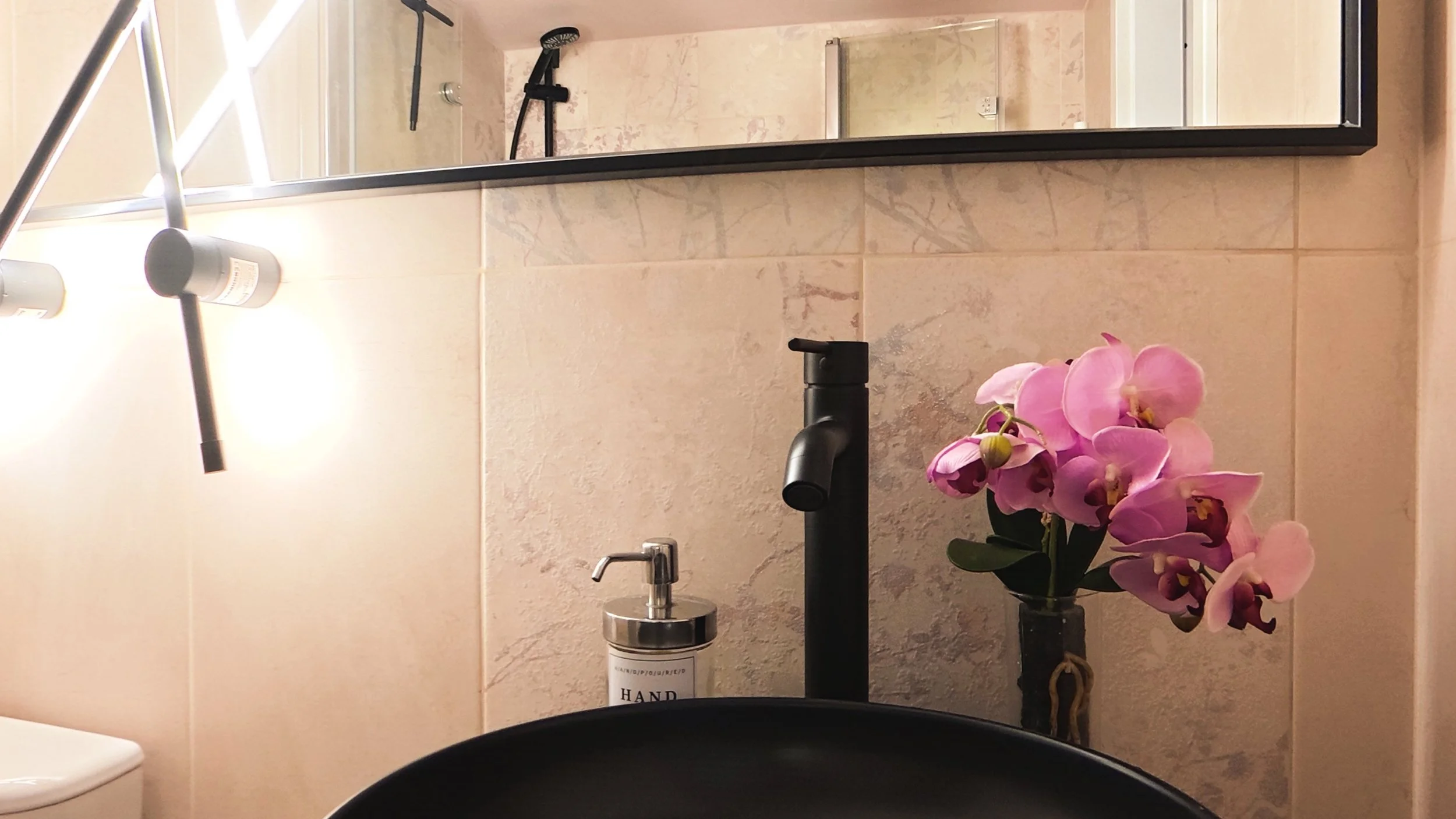 A bathroom sink with a black faucet, a mirror above, pink orchids in a black vase, and part of a toilet visible on the left. The wall behind features beige tiled walls and the reflection shows a showerhead.