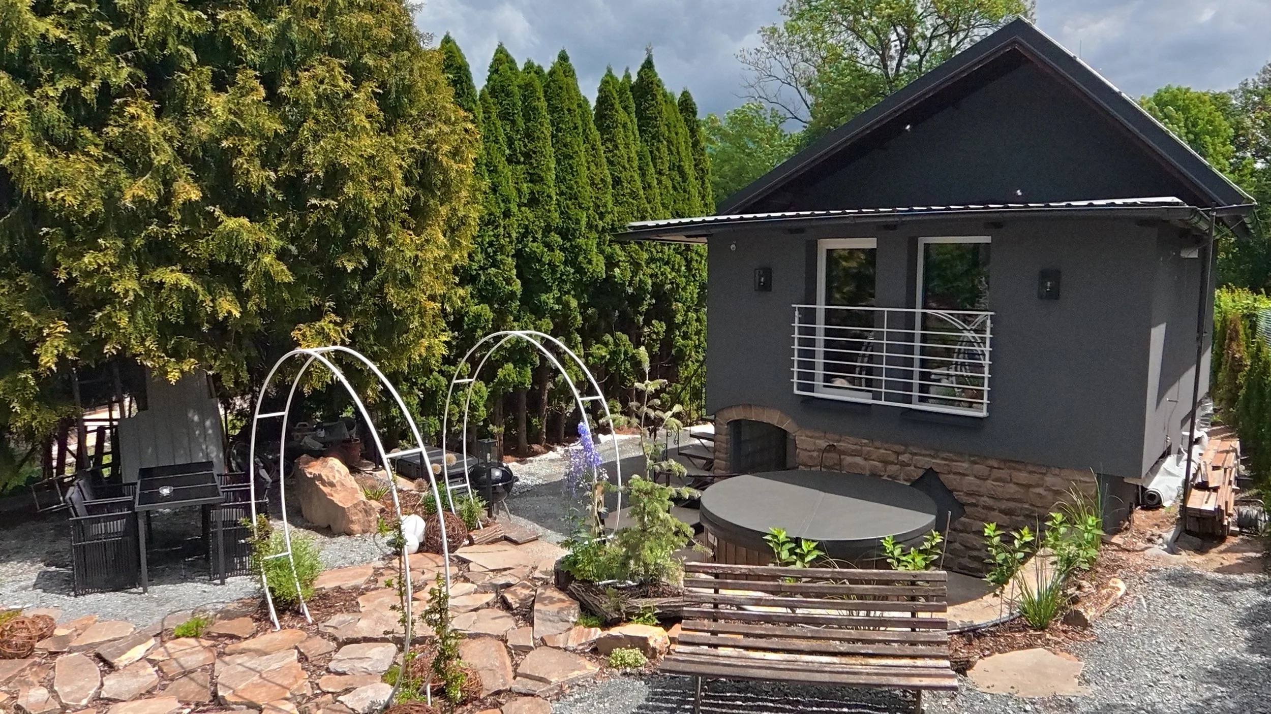 View of a backyard garden with outdoor seating, greenery, and a gray house with a balcony.