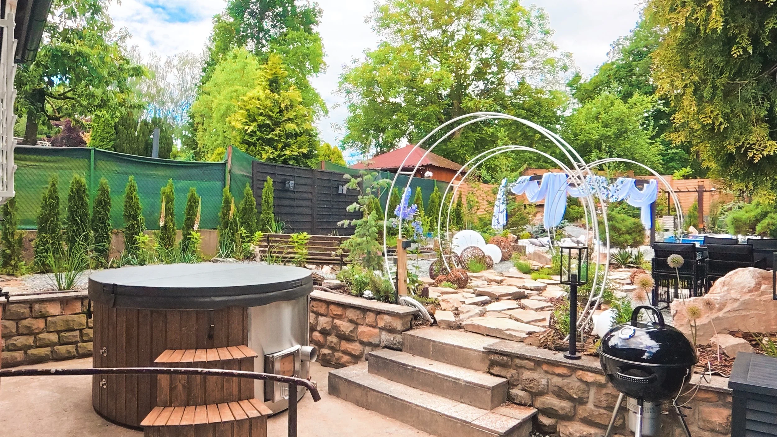 A backyard with stone steps, a black grill, a hot tub, and decorative arches with fabric, surrounded by greenery, trees, and garden plants.