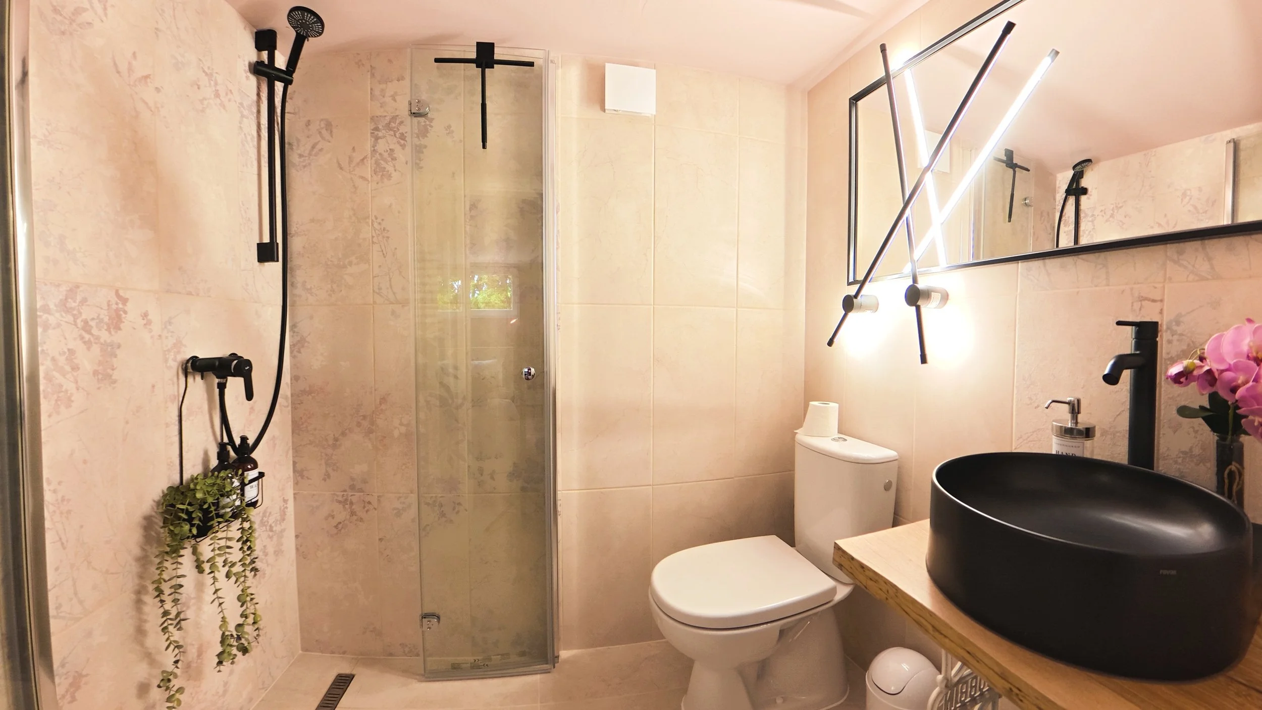 Modern bathroom with beige tiled walls and floor, a glass shower enclosure with black shower fixtures, a white toilet, a black sink on a wooden countertop, and a large mirror with contemporary lighting. Decor includes pink flowers and hanging plants.