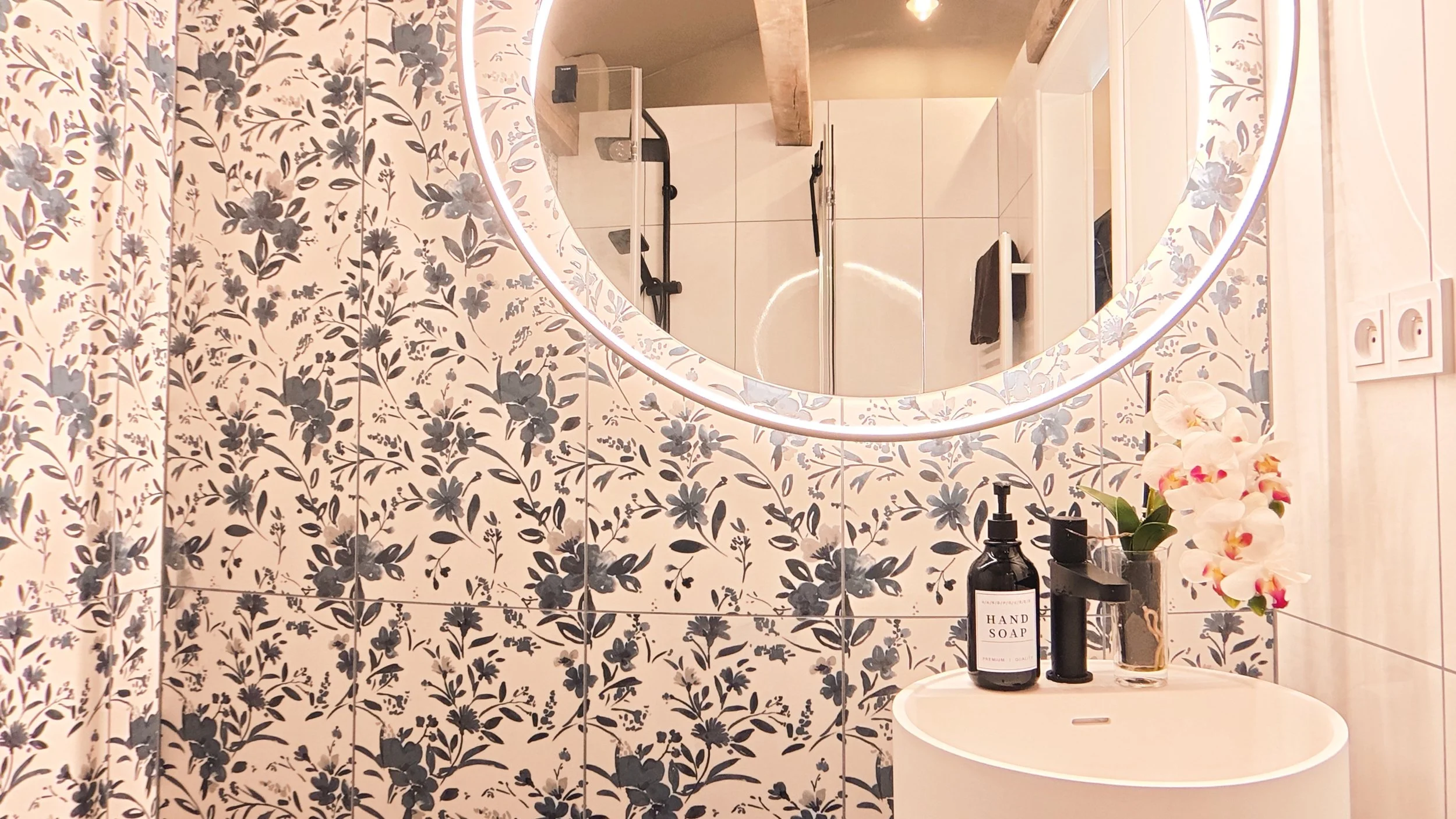 A bathroom with floral black and white patterned tiles, a round illuminated mirror, a white sink, and a small shelf with toiletries including a black bottle of hand soap and a glass vase with white and pink orchids.