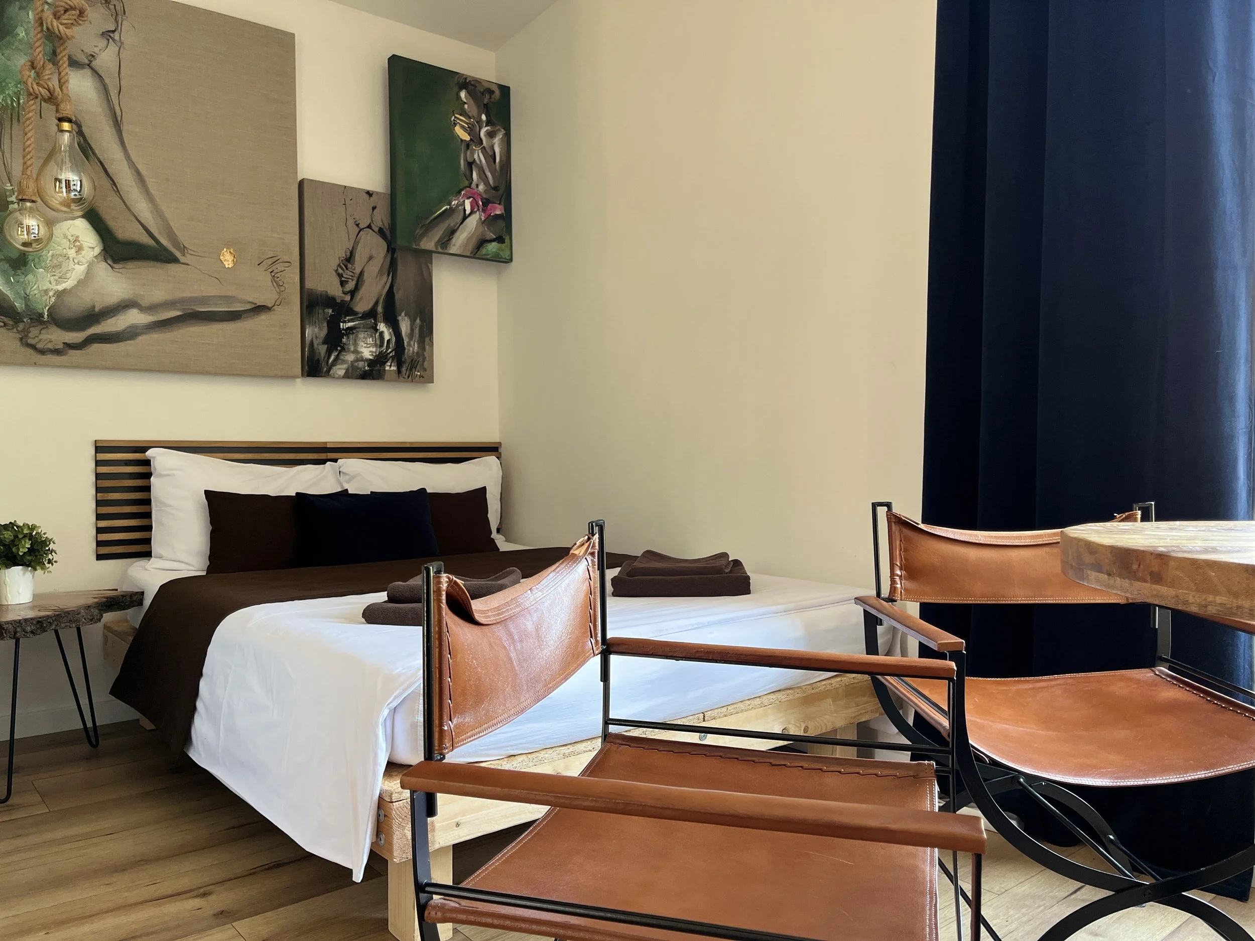 A bedroom with a bed that has a wooden slat headboard, white bedsheets, and pillows. There are three paintings on the wall above the bed, including a drawing of a woman and an abstract painting of a person. A small plant is on a side table, and a cluster of light bulbs hangs beside the paintings. A round wooden table and two leather chairs are in the foreground.