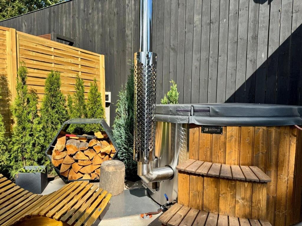 An outdoor hot tub with a wooden exterior, surrounded by potted plants, small trees, a wood storage rack, and a sauna stove, enclosed by a wooden privacy fence.