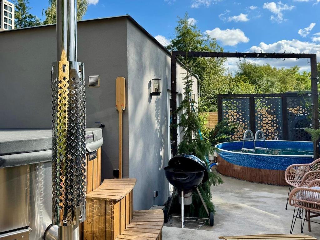 Backyard patio with above-ground pool, outdoor furniture, a grill, and a hot tub, surrounded by a wooden privacy fence and lush trees, on a sunny day.