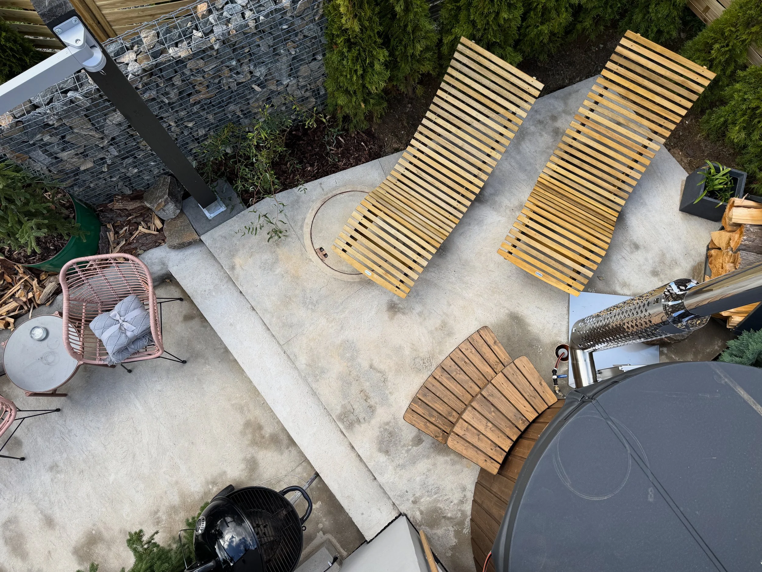 A backyard patio with two wooden lounge chairs on concrete, surrounded by green bushes, a small potted plant, and a grill.