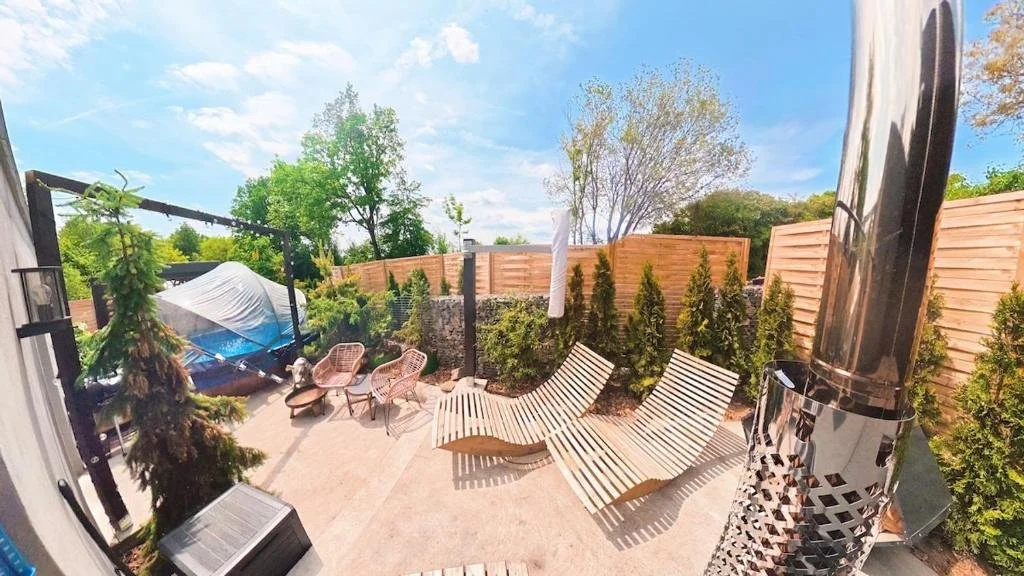 A backyard patio with wooden lounge chairs, wicker chairs, a covered hot tub, tall fence, and trees with a blue sky