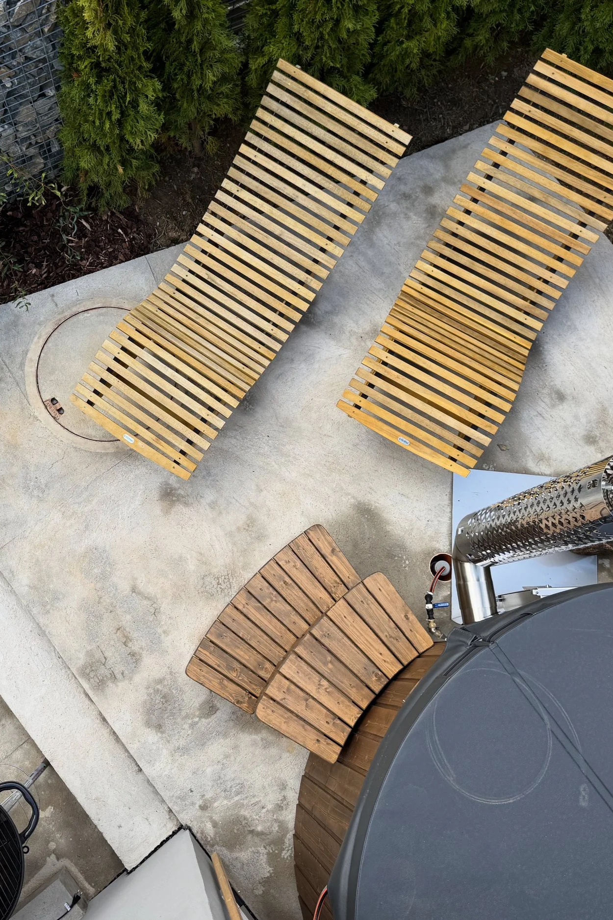 Two outdoor wooden lounge chairs on a concrete patio, with a third wooden piece nearby. There are green bushes along the edge of the patio, and a metal fence is partially visible in the background.
