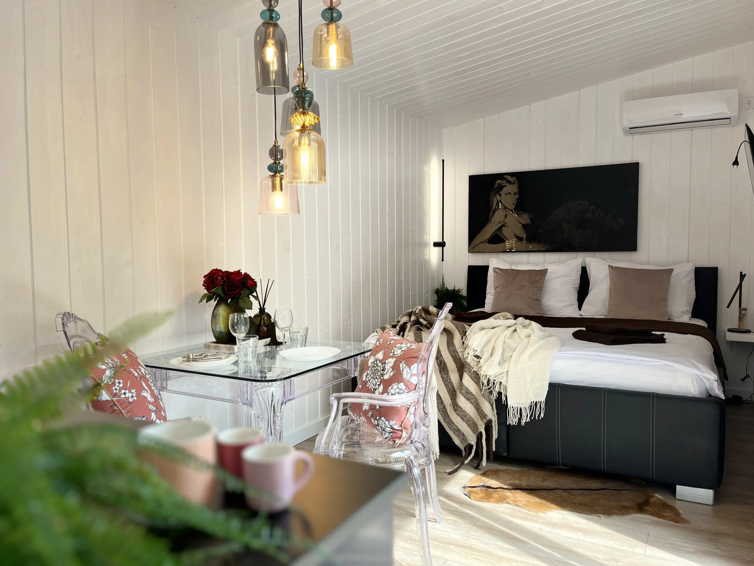 A bedroom with white shiplap walls and ceiling, a black portrait of a woman hanging above the bed, transparent chairs with floral pillows, a glass dining table set with wine glasses and a flower vase, and hanging pendant lights.