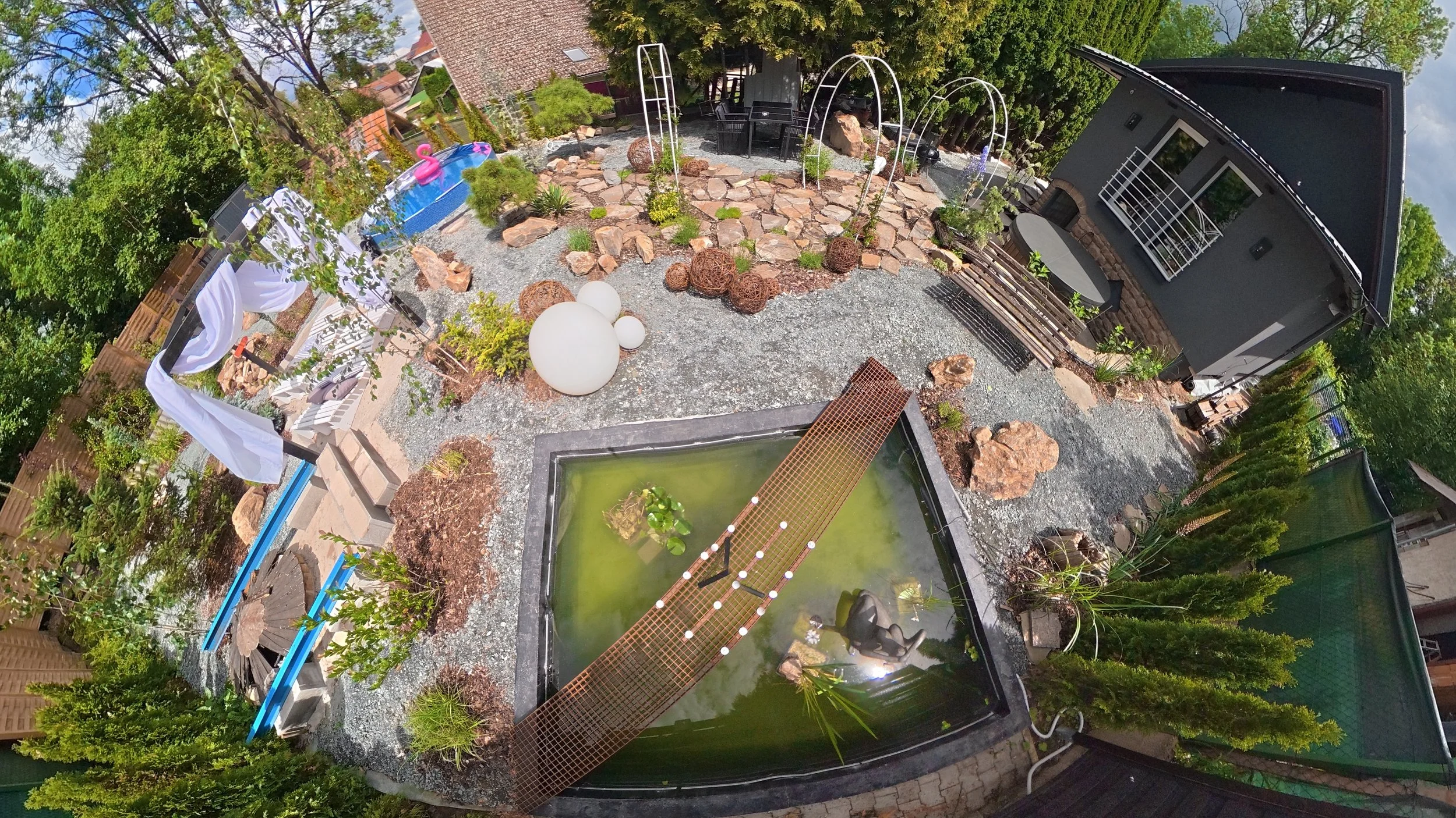 A backyard garden with a small pool with a pink flamingo float and a pond with a frog statue and green algae. There are decorative lights and various plants, rocks, and outdoor furniture.
