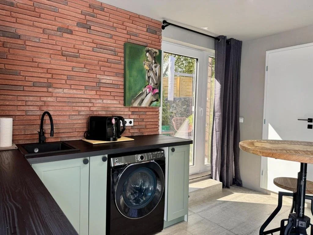A modern kitchen area with a black washing machine, a black faucet, a black coffee maker, and light green lower cabinets. There is a painting of a woman on a red brick wall, a sliding glass door with open curtains leading outside, and a wooden round table with a black base.
