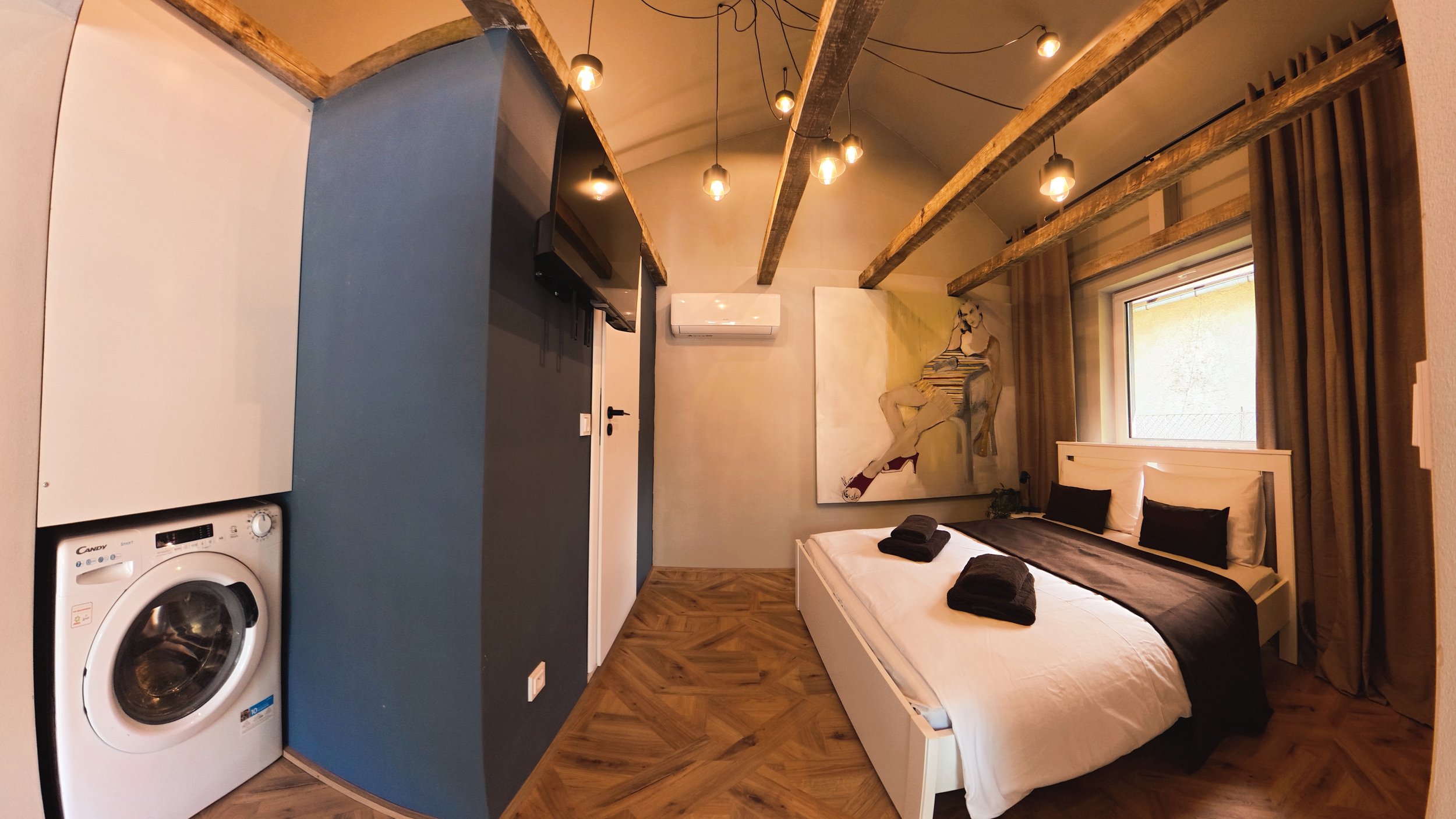 A bedroom with a white bed, black pillows, and neatly folded black towels on top. There is a large window with beige curtains, a painting of a woman hanging on the wall, and exposed wooden beams on the ceiling. A wall-mounted air conditioning unit is visible, and there is a washing machine in the corner.