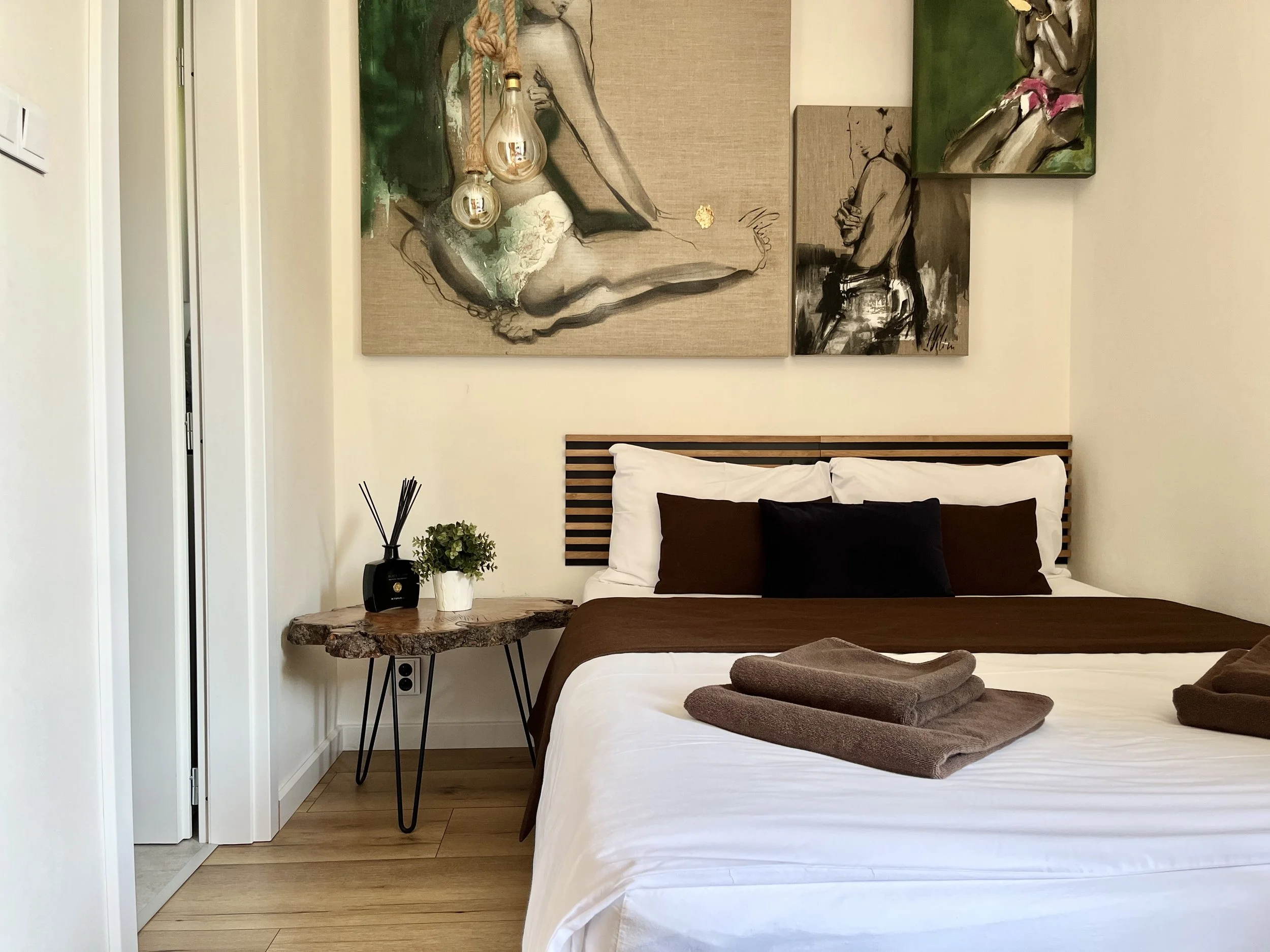 A modern bedroom with a bed with white and brown pillows, wooden headboard, and folded towels on the bed. A side table with a black vase and green plant, abstract art on the wall, and a rustic wooden table are visible.