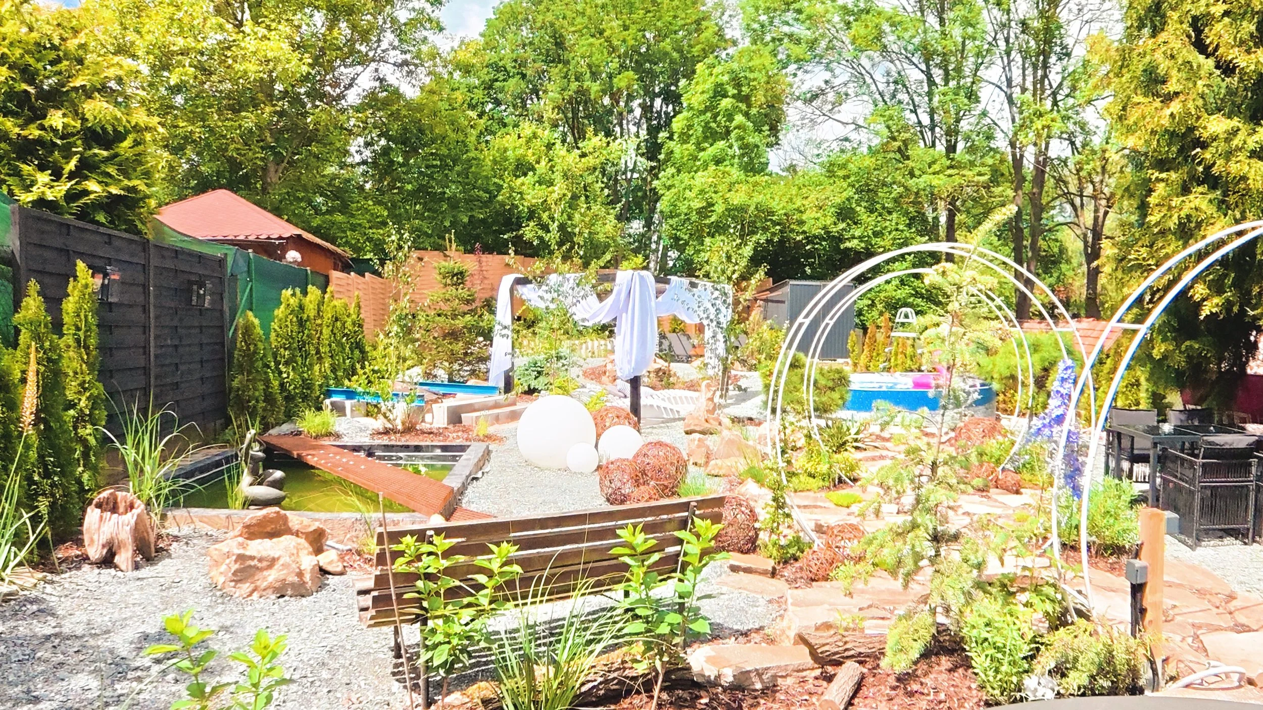 A lush garden with various plants, trees, and decorative elements including white spherical lights and geometric wire structures under a bright blue sky.