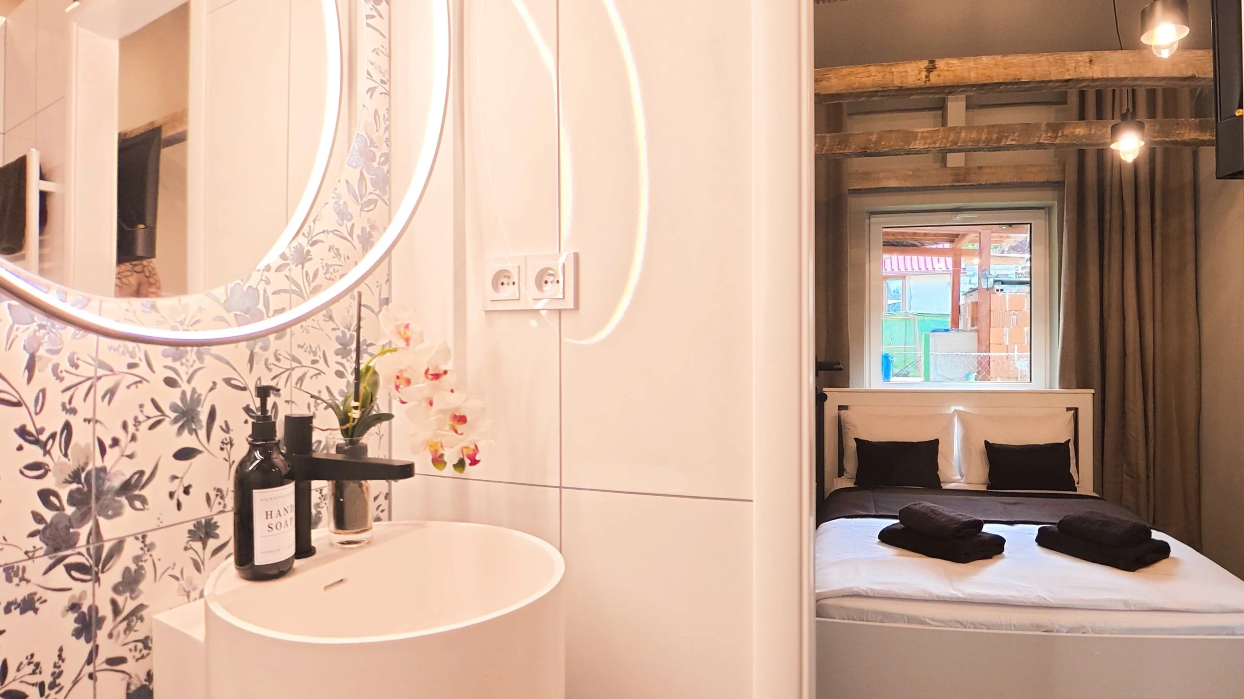 A bathroom with a round mirror, floral wallpaper, a sink with soap, and a small plant. Adjacent is a bedroom with a bed, pillows, window, and wooden beams.