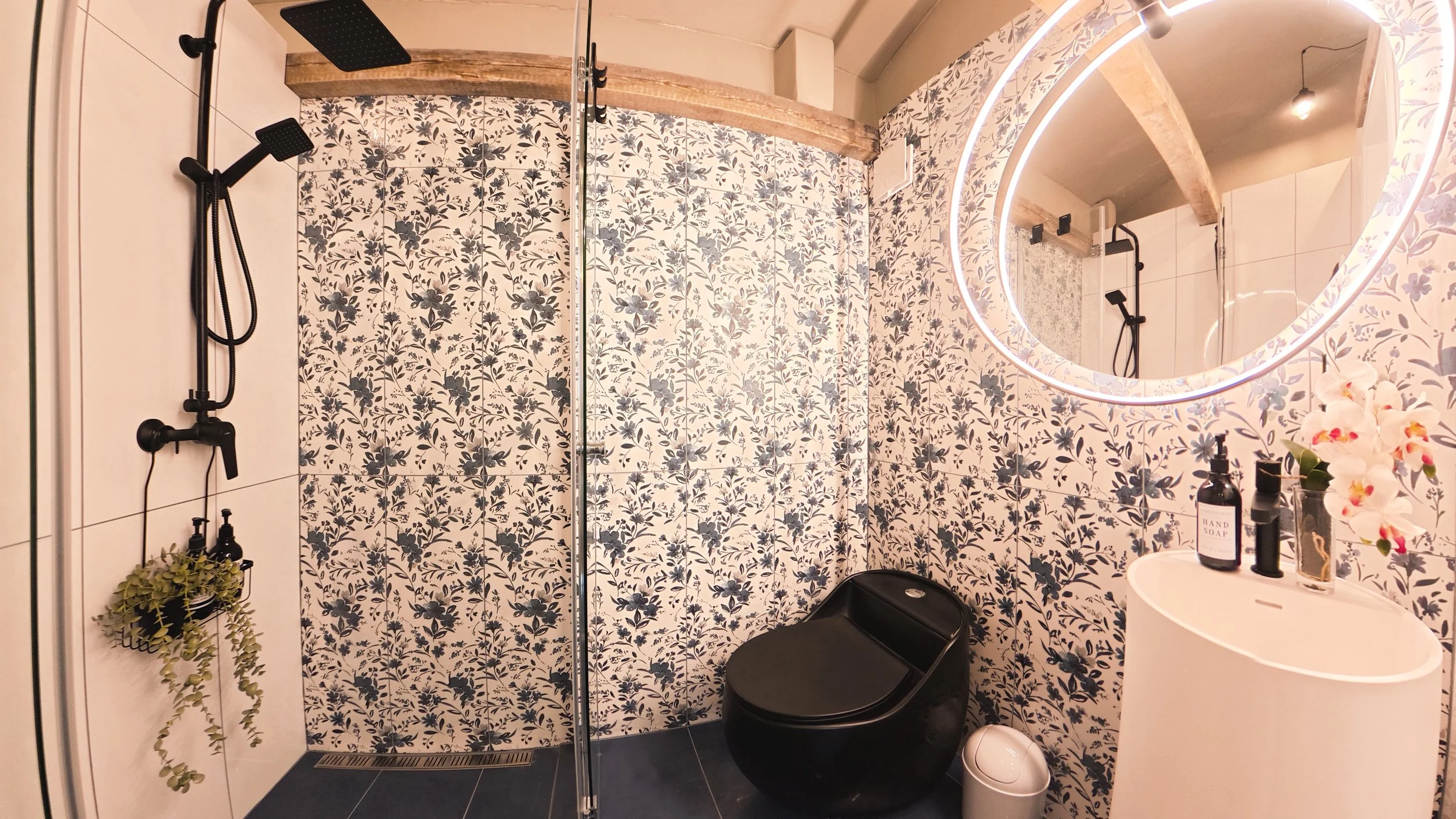 Modern bathroom with floral patterned tiles, a black shower with rainfall and handheld showerheads, a round mirror with LED lighting, a white sink counter with hand soap and flowers, and various toiletries and plants.