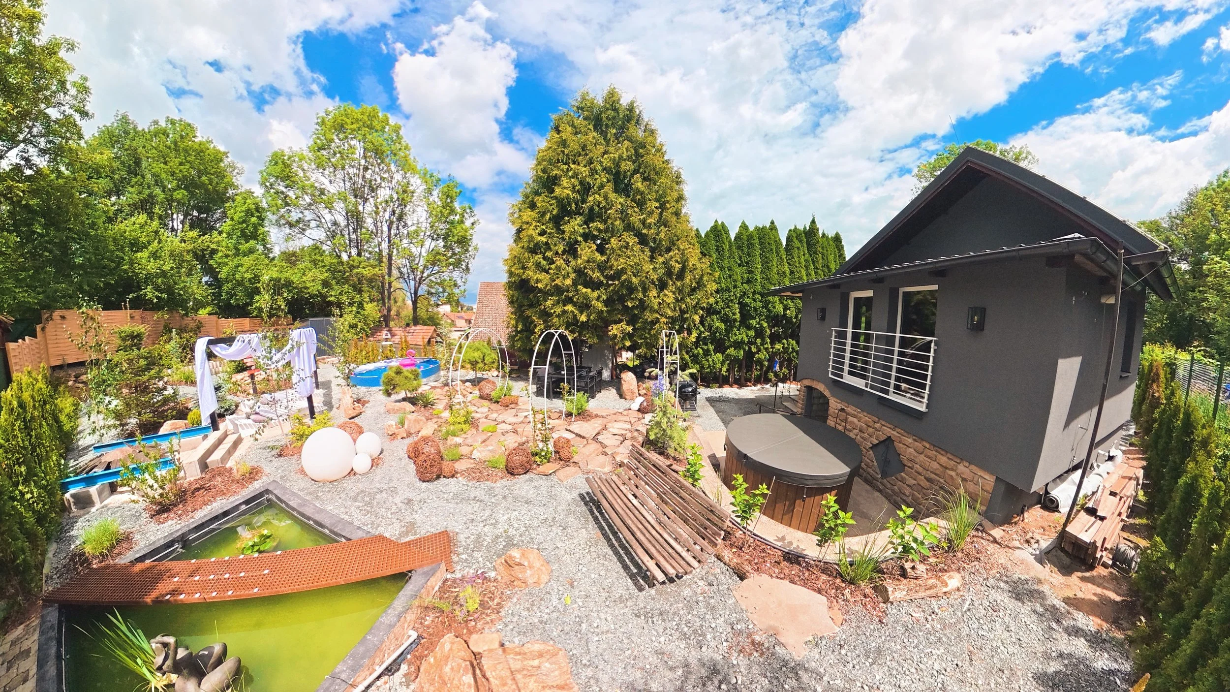 A backyard with trees, a gray house, a small pond, and a blue inflatable kiddie pool. There are decorative plants, rocks, and outdoor furniture visible under a partly cloudy sky.