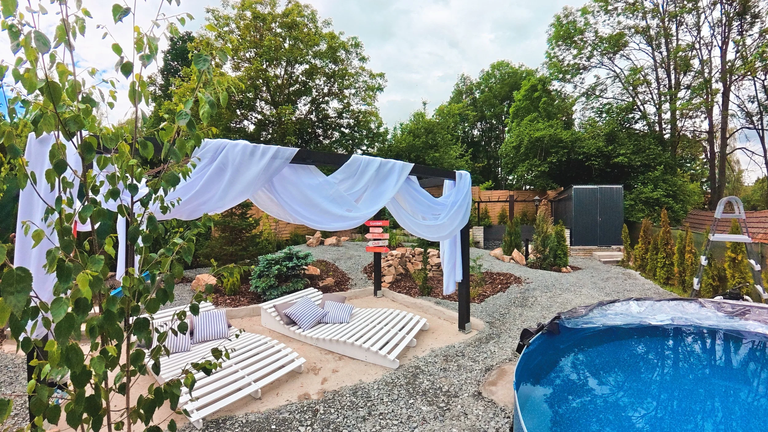 A backyard patio with white lounge chairs, pillows, a pergola draped with a white fabric, small bushes, and a signpost, surrounded by trees and a wooden fence.
