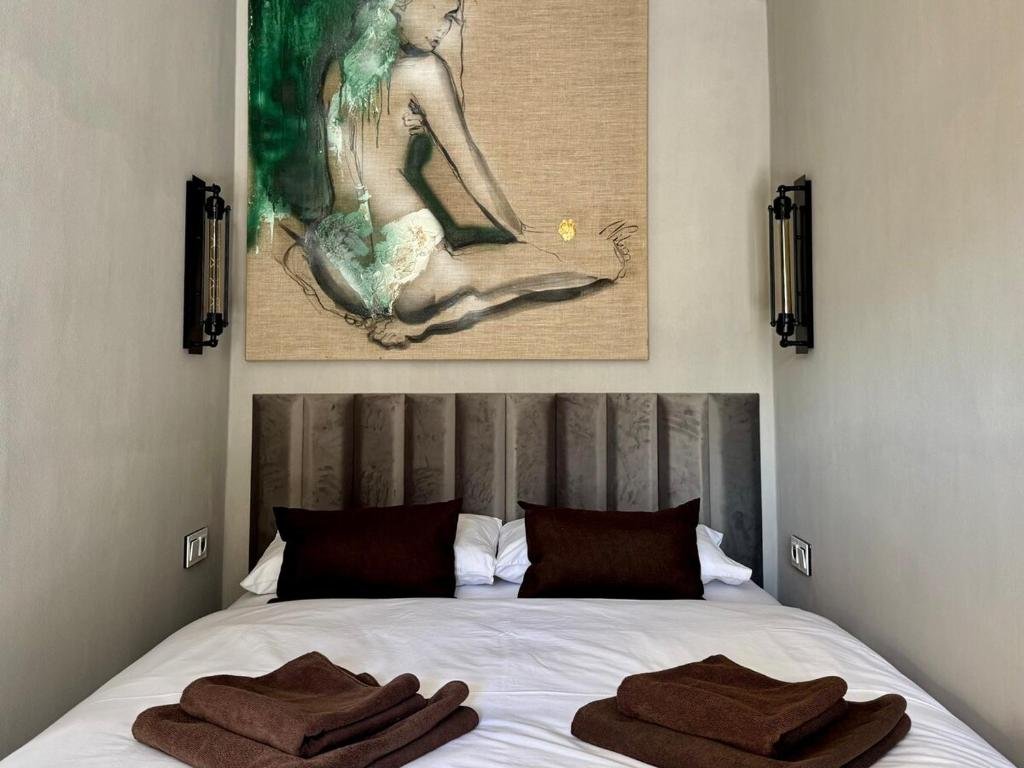A neatly made bed with brown pillows and folded towels on top, set against a padded headboard with artwork above it, in a modern bedroom with wall-mounted lights.