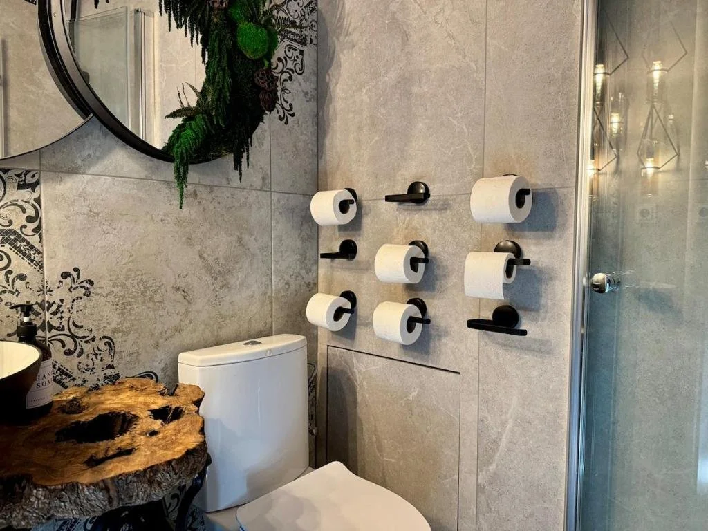 A bathroom with grey tiled walls, a toilet, a small sink with a black soap dispenser, a mirror, and a unique wooden countertop. A wall-mounted holder with six rolls of toilet paper is visible.