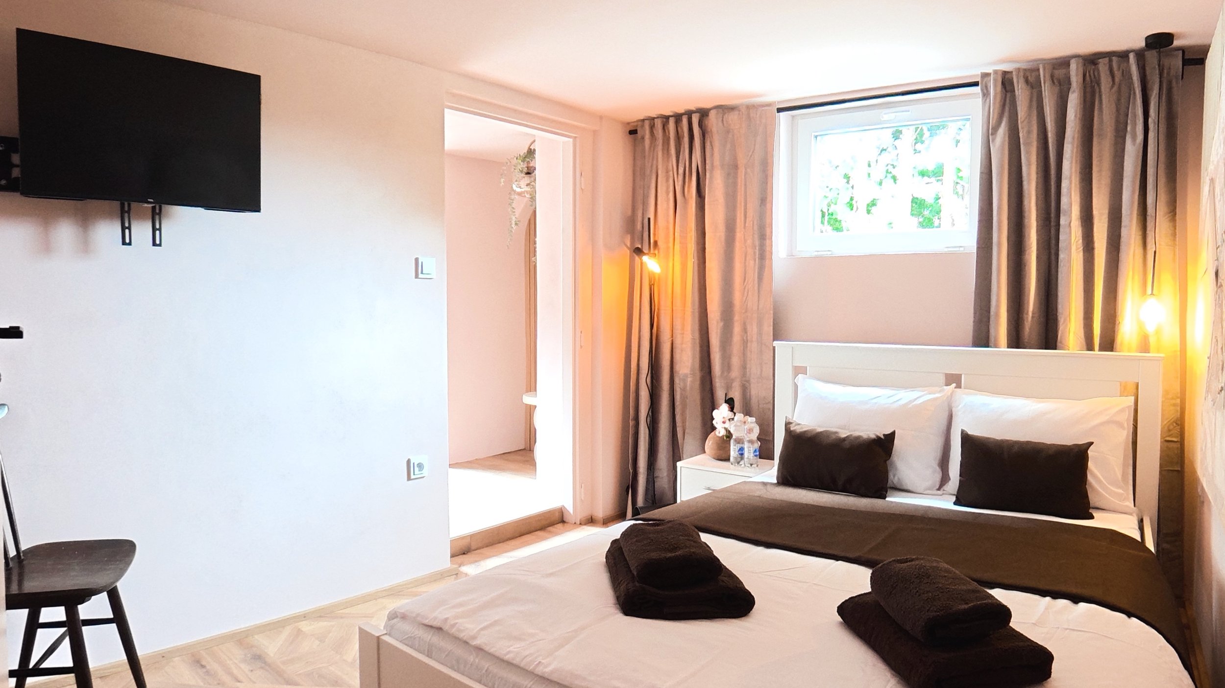 A cozy bedroom with a large bed covered in white and brown bedding, black pillows, and neatly folded brown towels. There is a small white nightstand with bottled water and flowers, beige curtains by a window, and warm lighting from two lamps. A flat-screen TV is mounted on the wall.