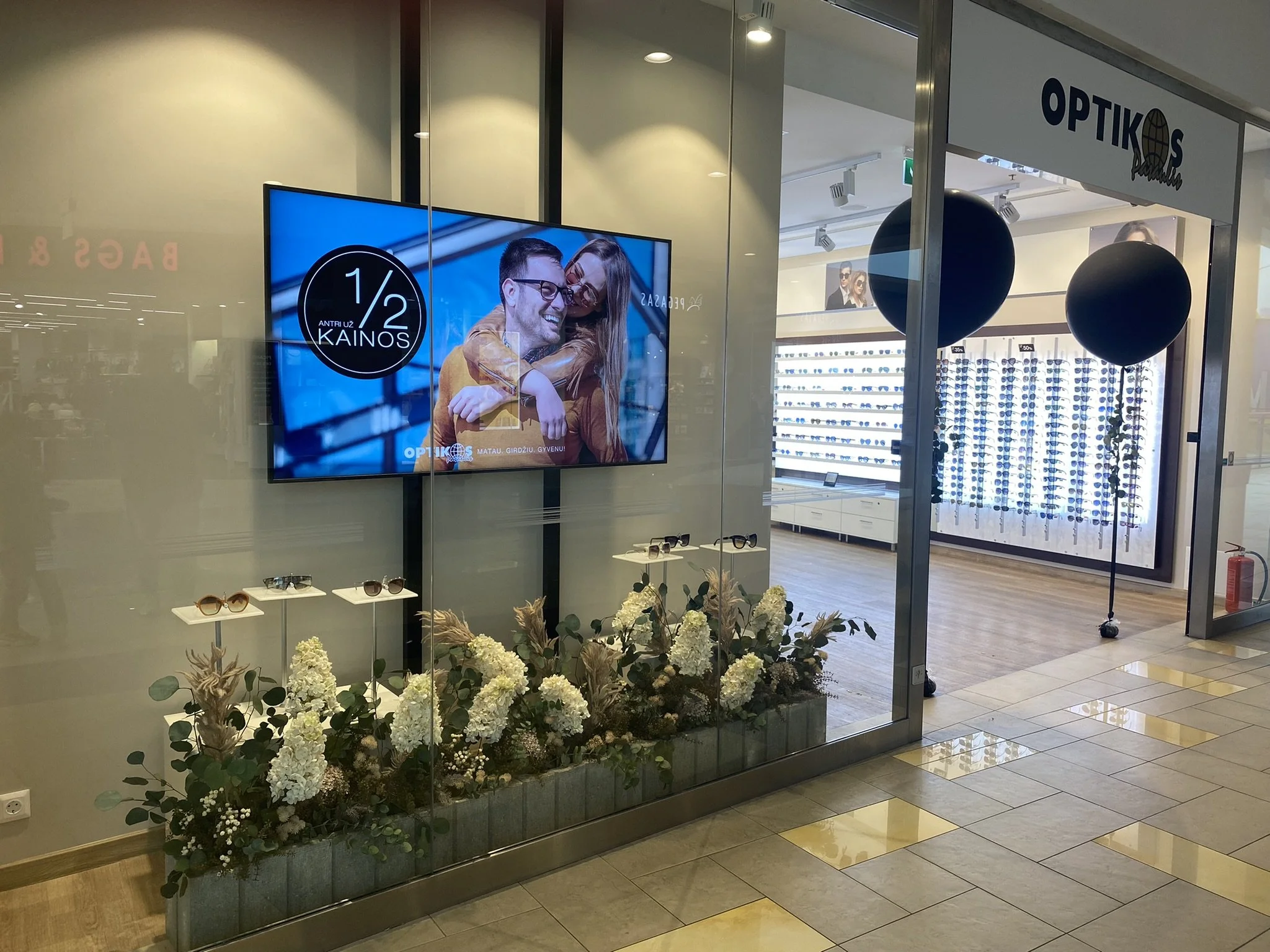 A storefront display window for optical glasses with floral decoration at the base, a digital advertisement showing a smiling man and woman wearing glasses, and additional eyeglasses on small white tables inside the store.