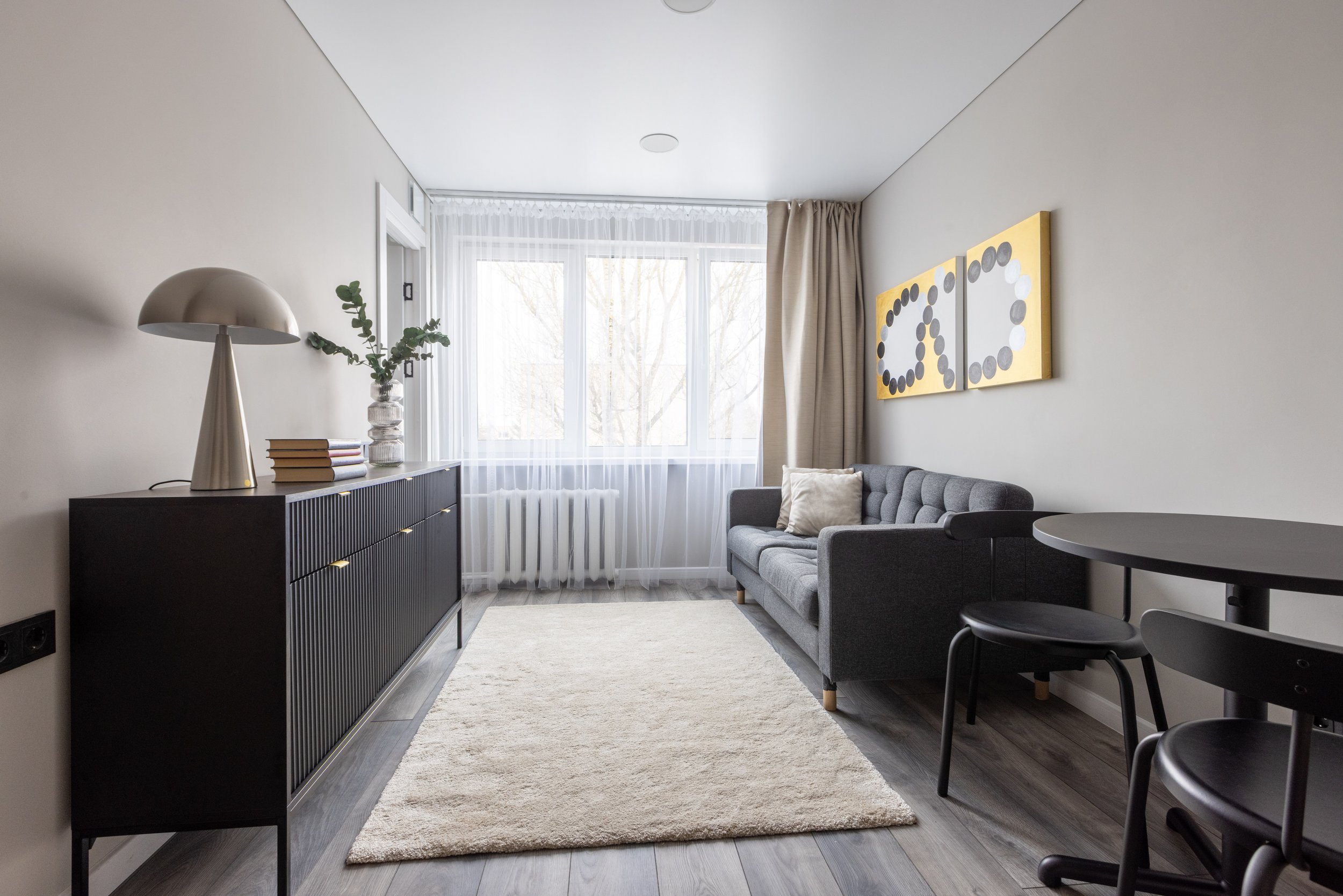 Interior of a living room with a gray sofa, a black sideboard with a lamp and books, a vase with green foliage, large windows with sheer curtains, and modern wall art with yellow backgrounds and black and white circles.