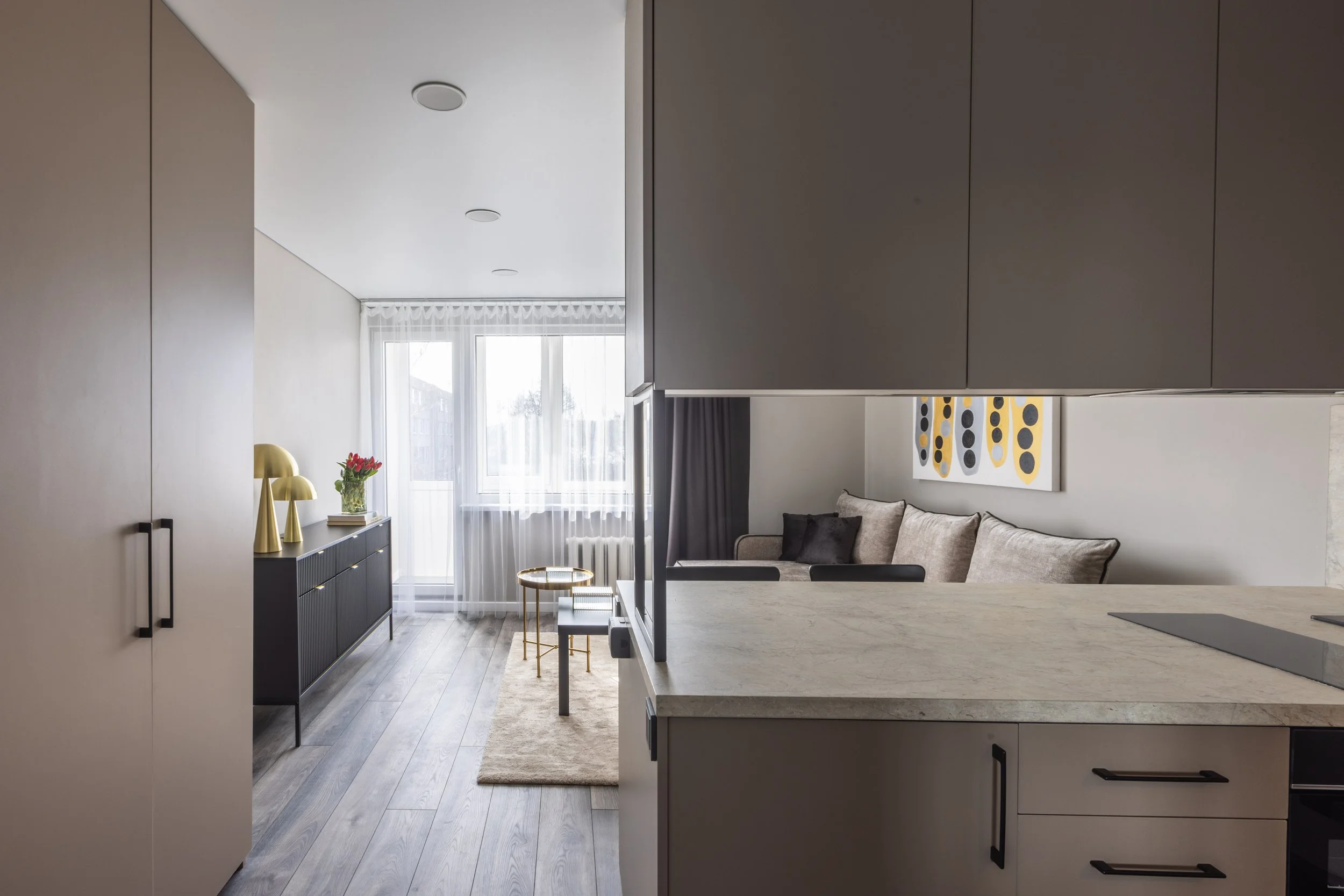 Modern open-plan living room and kitchen with large windows, grey cabinets, artwork, and decorative lamps.