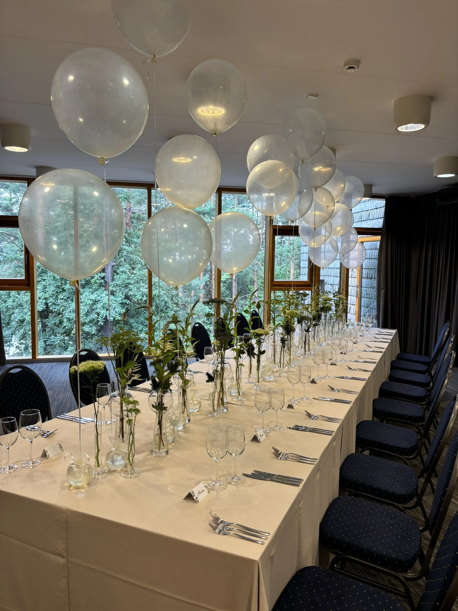 Elegant banquet table set for a formal event, decorated with vases of flowers, surrounded by dark blue chairs with polka dots, and ceiling of the room with balloons and lighting fixtures, overlooking a window with greenery outside.