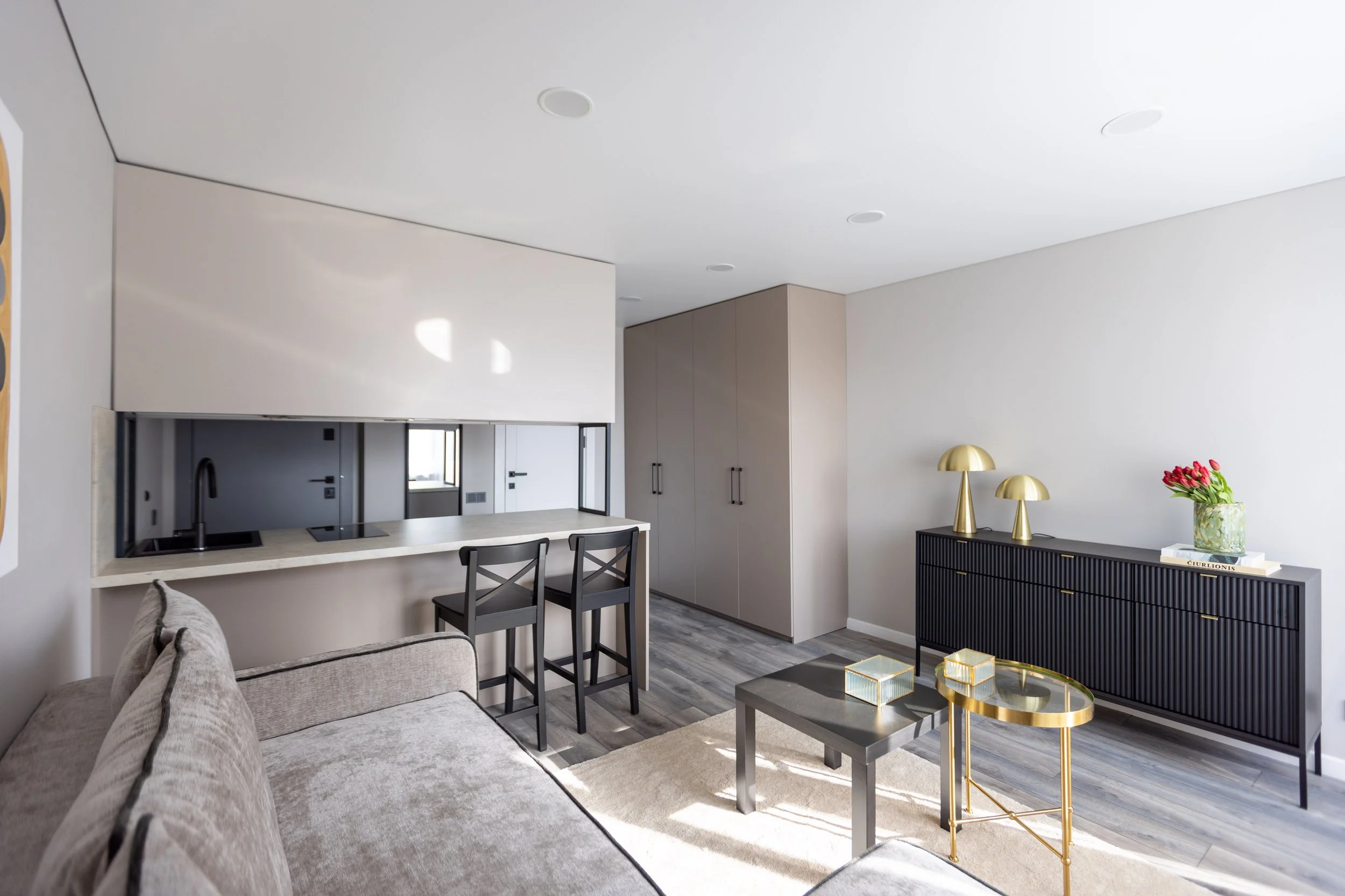 Modern living room with a beige sofa, black chairs at a kitchen counter, and a black console with gold lamps and a vase of pink flowers on top.