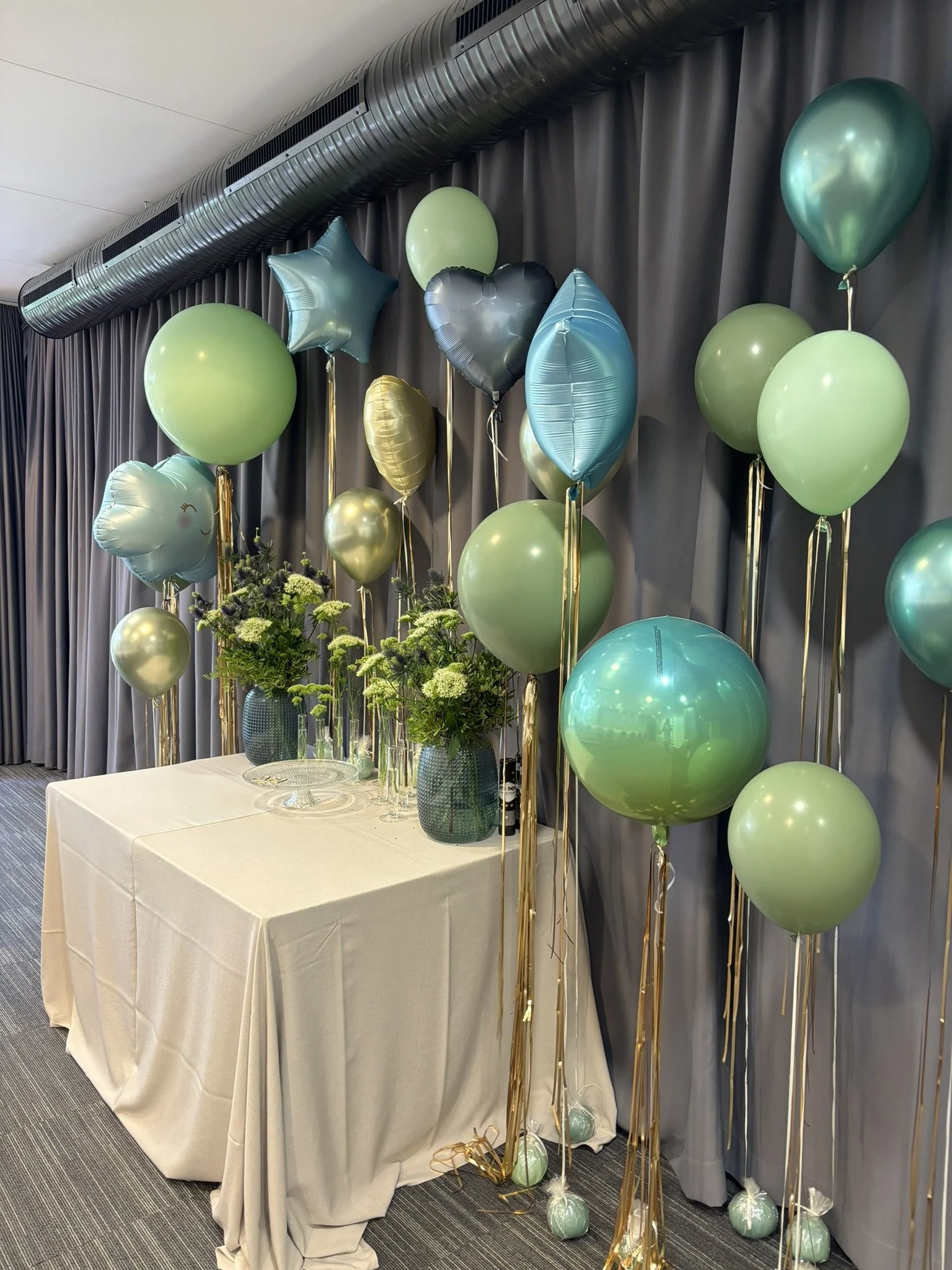A decorated party display with green and blue balloons, including star and heart-shaped balloons, arranged behind a table with floral arrangements in vases, against a dark curtain backdrop.