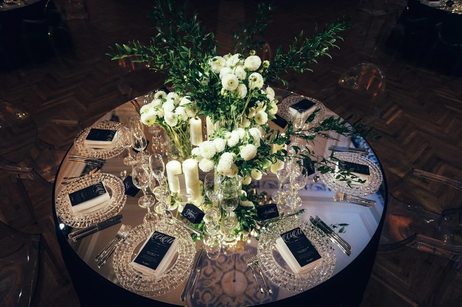 Elegant round table setup with a large floral centerpiece of white flowers and greenery, surrounded by candles, glassware, plates, and black menus, with transparent chairs and wooden flooring in the background.