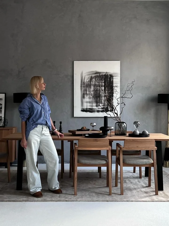 A woman with blonde hair wearing a blue striped shirt and white pants is standing beside a wooden dining table in a modern, minimalist room. The table is decorated with black and white artistic vases, bowls, and a branch. There is abstract black and 